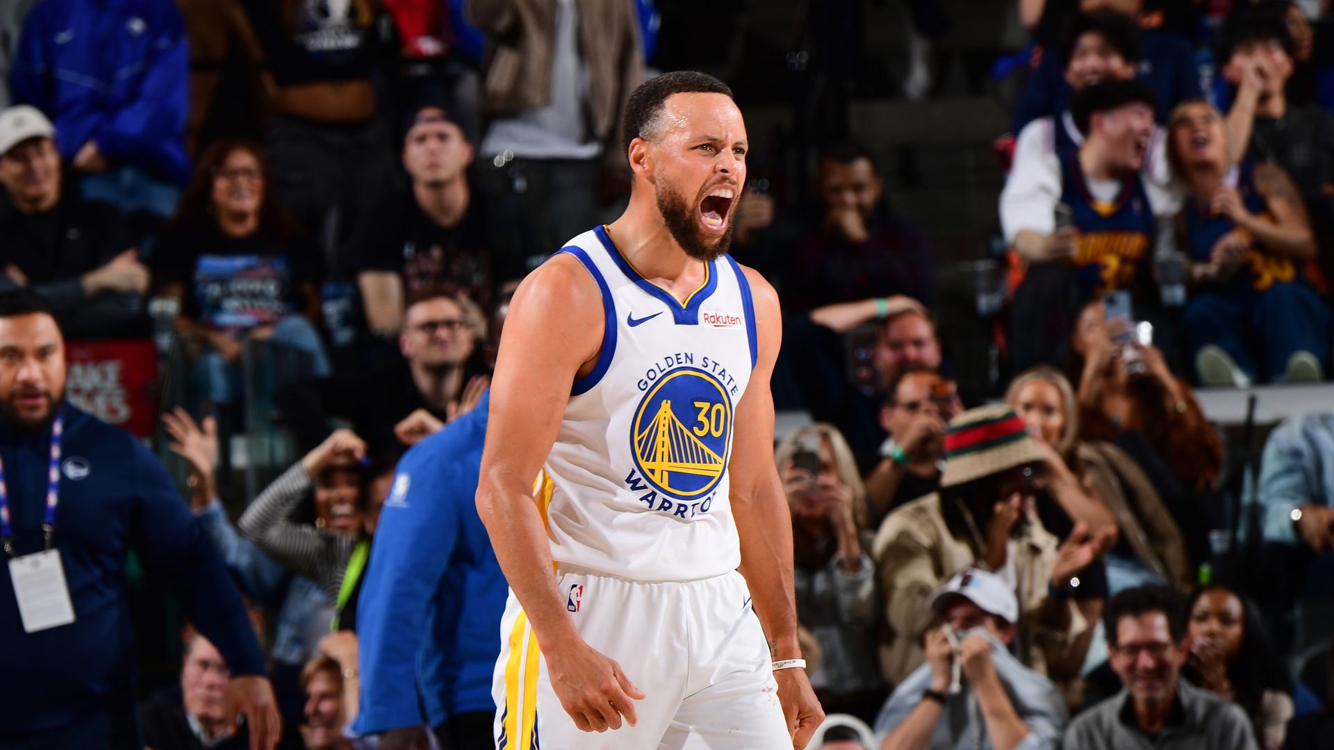 Golden State Warriors guard in a white jersey with blue and yellow trim, #30, roars on the court as fans cheer wildly in the arena behind him.