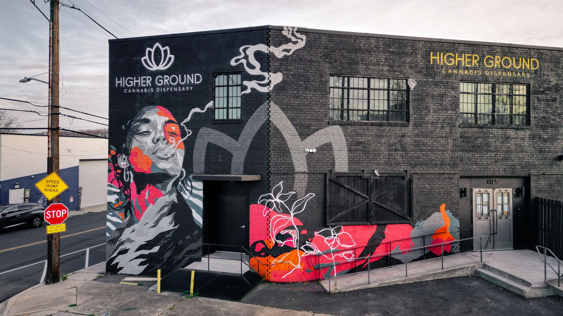 Street view of a gray brick building covered in a colorful mural; the sign reads "Higher Ground Cannabis Dispensary" in gold letters, with a utility pole, power lines, and a stop sign nearby.