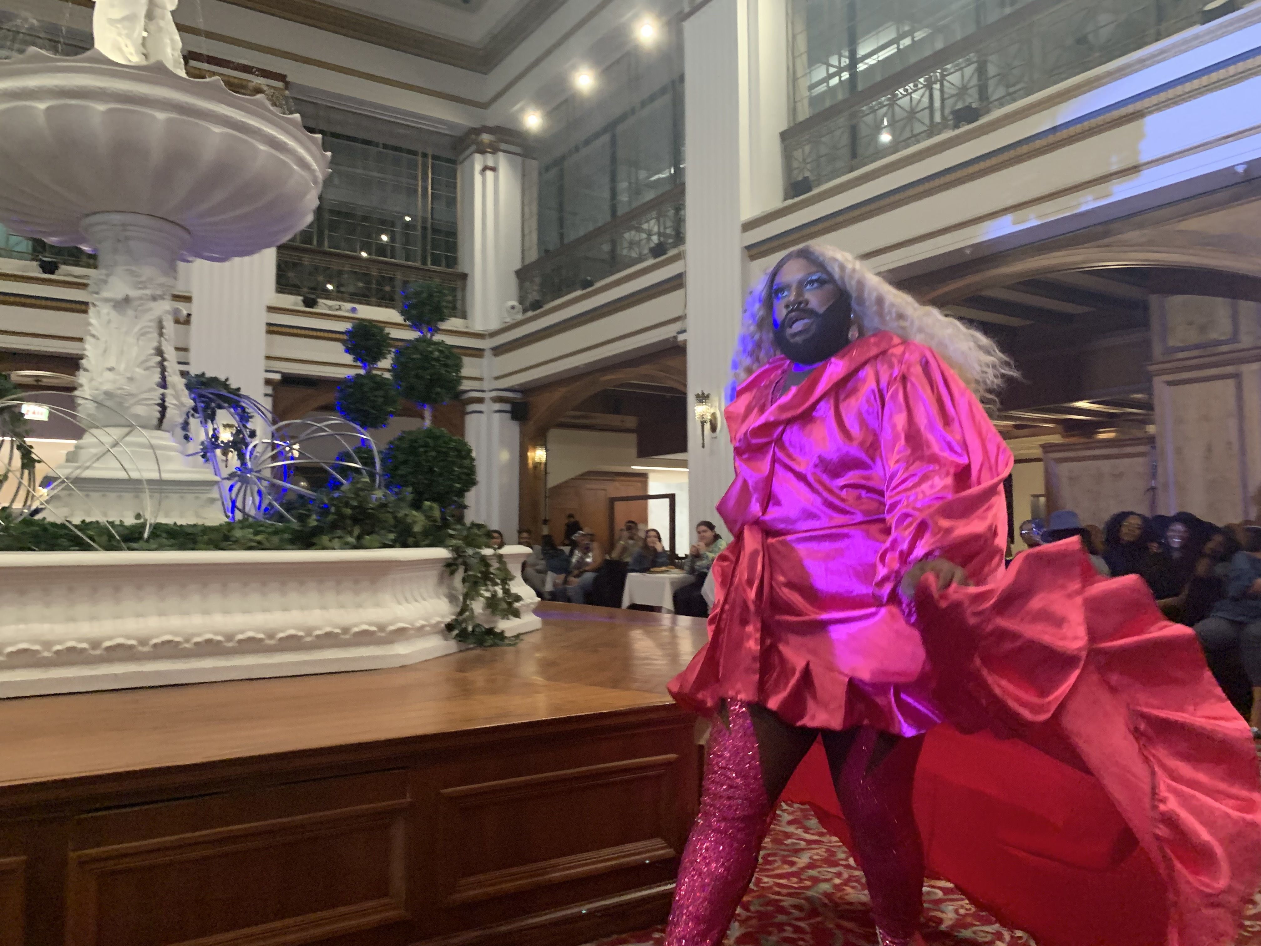 A drag performer in a bright pink dress and tights performs in front of the Walnut Room's fountain.