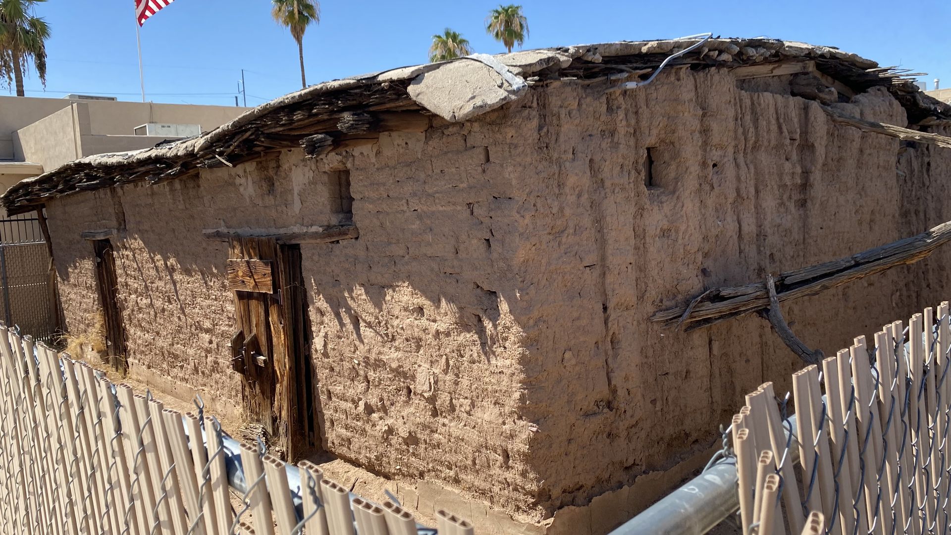 A crumbling old adobe house behind a chain link fence. 