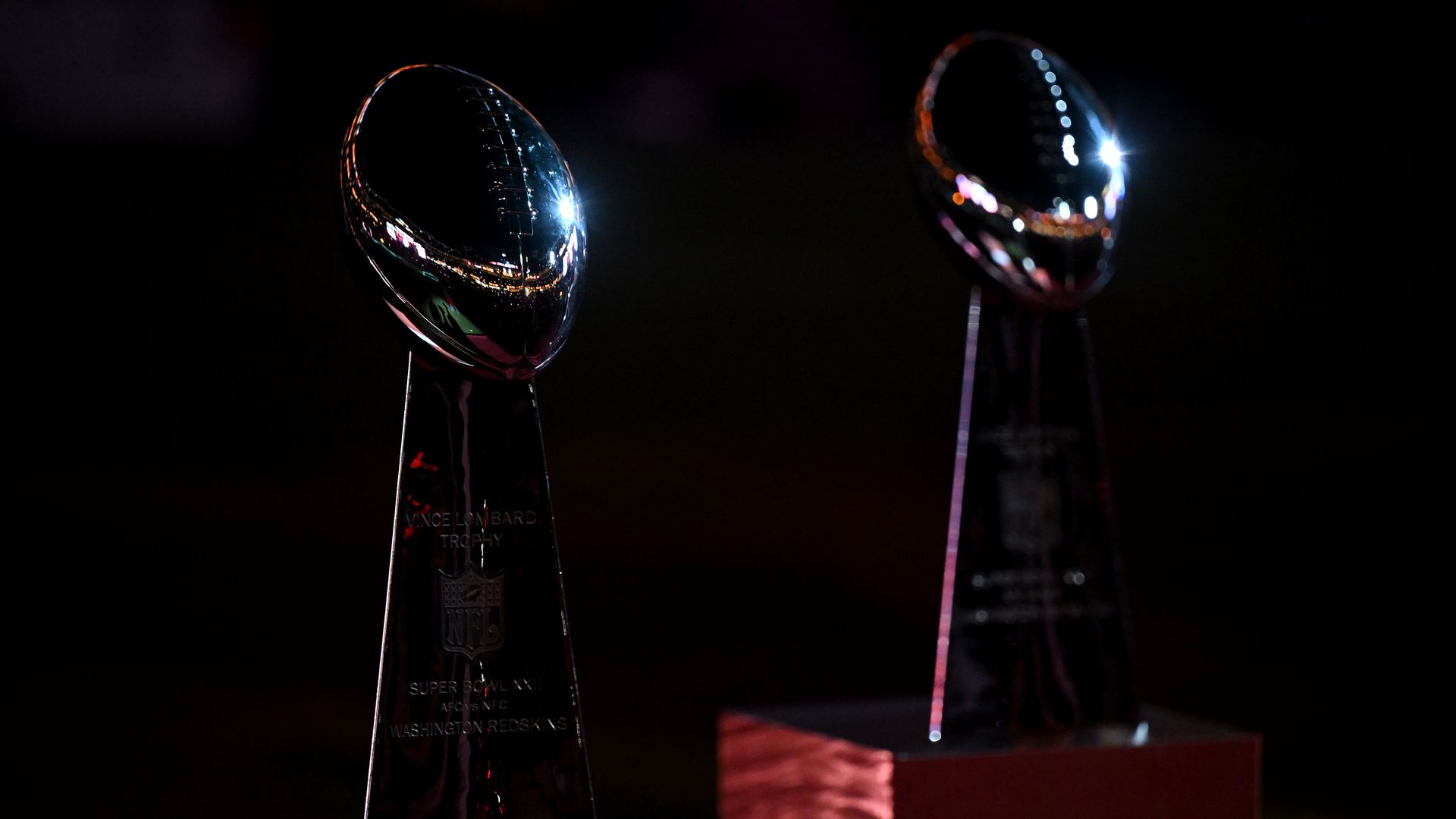 Two shiny Vince Lombardi trophies on dark pedestals in a dim setting; left one shows engravings, right one reflects colorful lights.