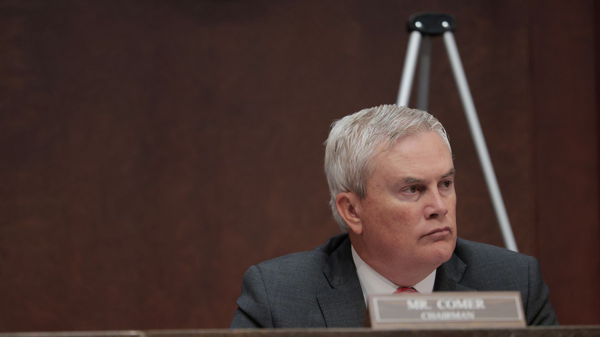 An older man with white hair in a dark suit and red-striped tie sits at a desk with a nameplate reading "Mr. Comer Chairman" against a brown background.