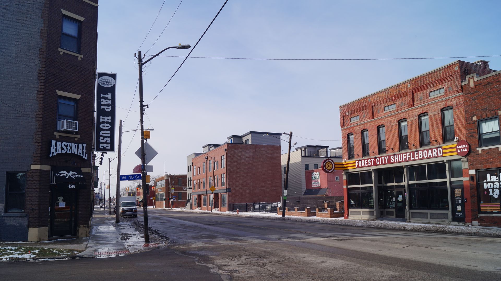 Empty street intersection with signs for Arsenal Tap House and Forest City Shuffleboard Bar in brick buildings under a cloudy sky, light snow on sidewalks.