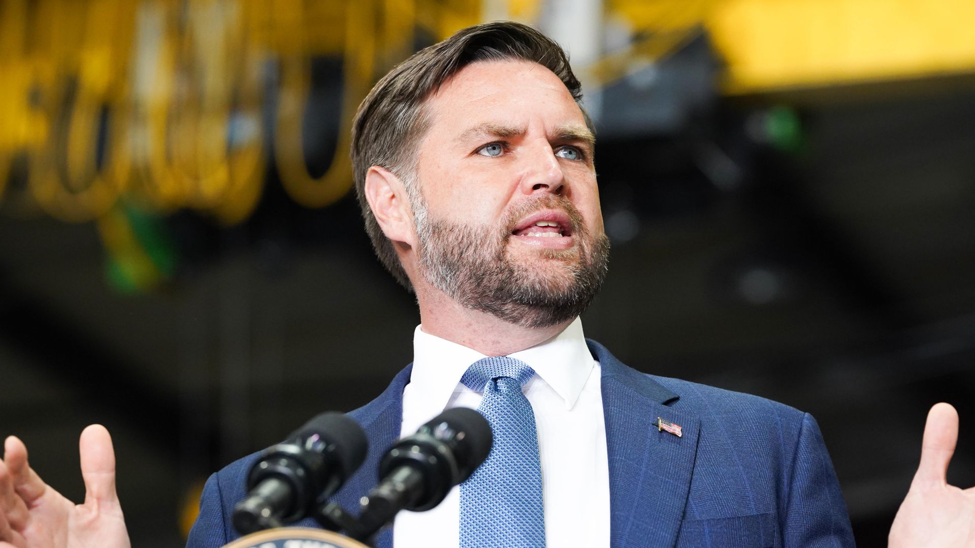JD Vance speaks into a microphone while holding up his hands and wearing a blue suit.