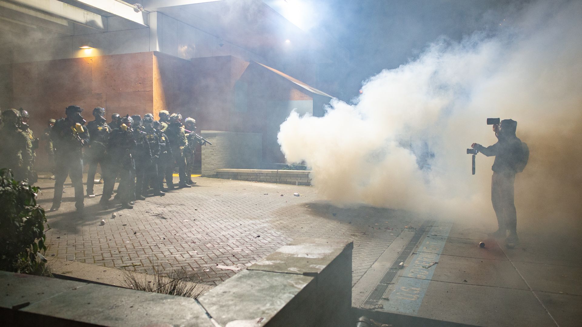 Nighttime urban scene: riot police in full gear form a line as thick smoke fills the air; a photographer with a camera stands to the right, with bright lights and a building behind.