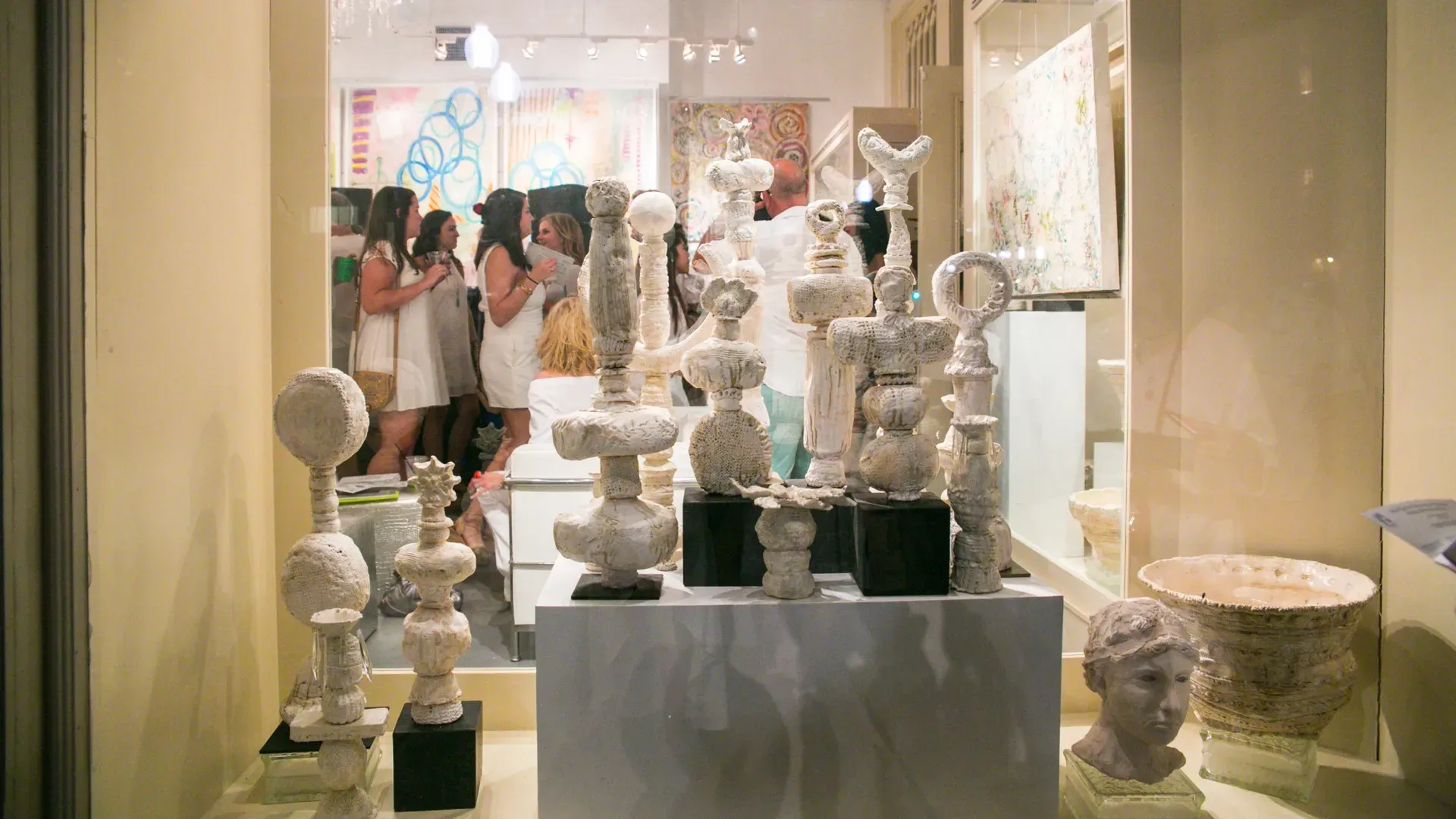 Art gallery window display featuring white abstract sculptures on pedestals, with people socializing and colorful paintings visible inside the well-lit space.