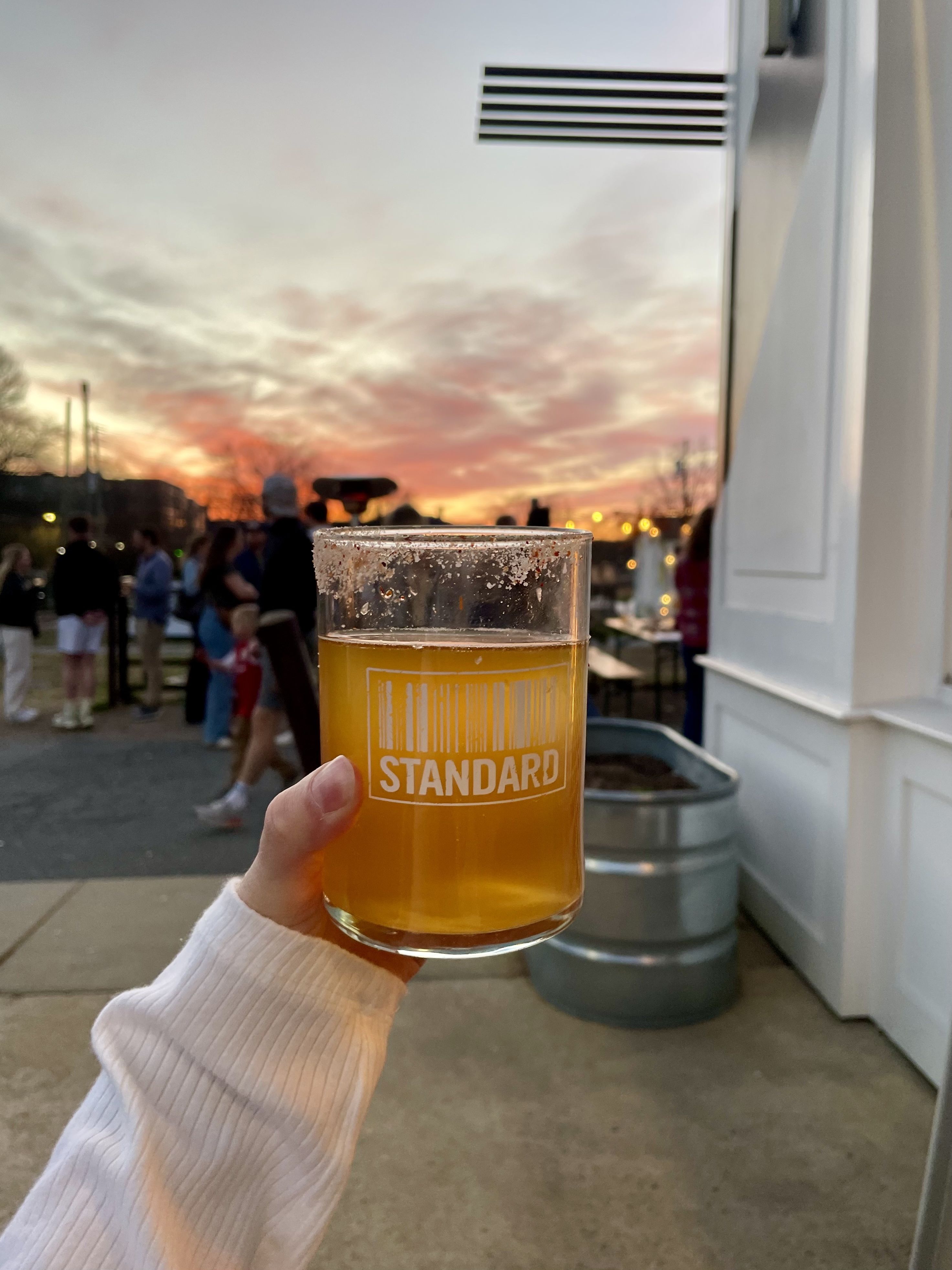 Hand holding a glass of amber beer with a salted rim labeled "STANDARD" against a sunset sky with blurred people and outdoor setting in the background.