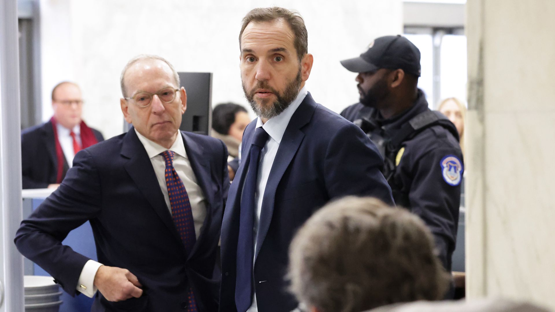 Bearded man in a navy suit and blue tie stands center, looking at the camera; to his left, an older man in a dark suit with glasses; a security guard in black with a patch stands in the background.