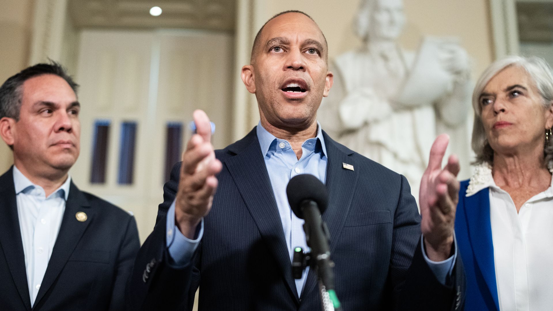 Center: a man in a dark suit and blue shirt speaks at a microphone, gesturing with hands. On both sides, a man and a woman listen as a marble bust stands in the beige background.