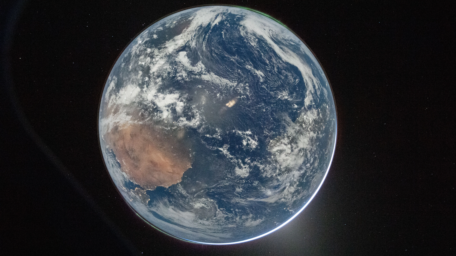 Earth viewed from space, showing Africa in the lower left with brown deserts, white cloud swirls over blue oceans, a thin glowing limb along the edge, and a small bright object in the dark background.