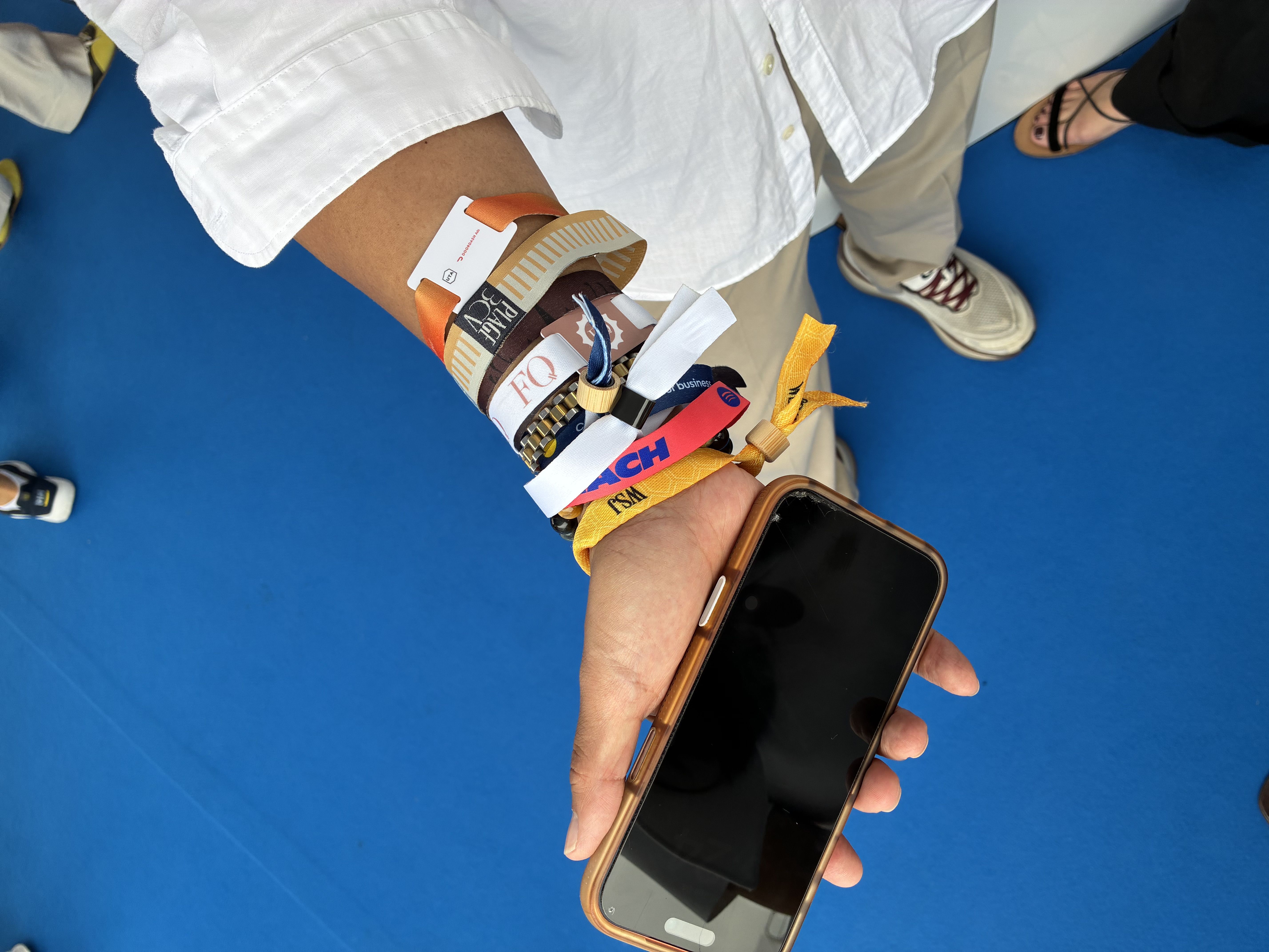 Close-up of a forearm wrapped in multiple colorful wristbands over a white shirt sleeve; a smartphone is held in the other hand, with a bright blue floor in the background.