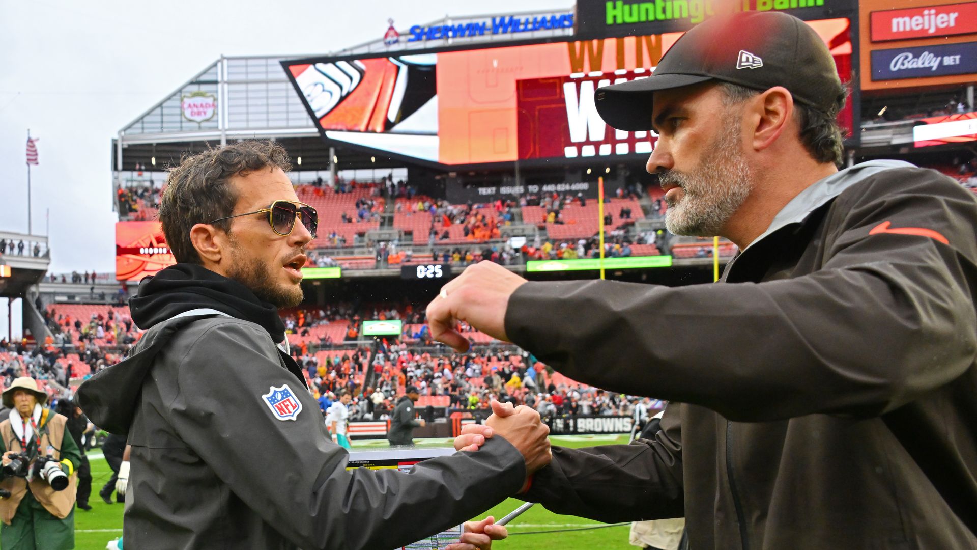 Two men in black NFL jackets fist bump on a football field with a stadium crowd and scoreboard in the background. One man wears sunglasses, the other a black cap.