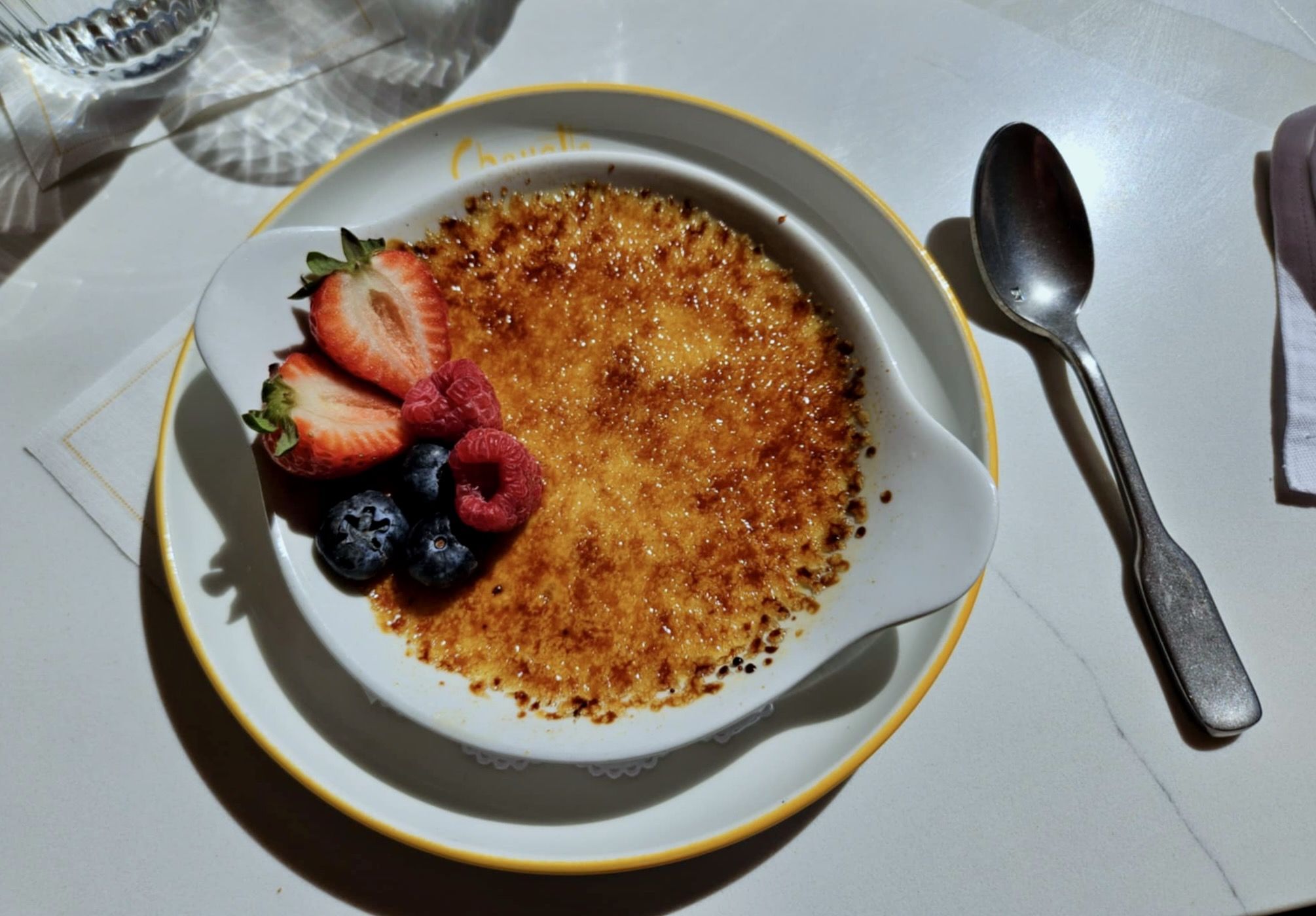 Caramelized crème brûlée in a white dish with sliced strawberries, raspberries, and blueberries on a plate with a yellow rim beside a metal spoon on a white table.