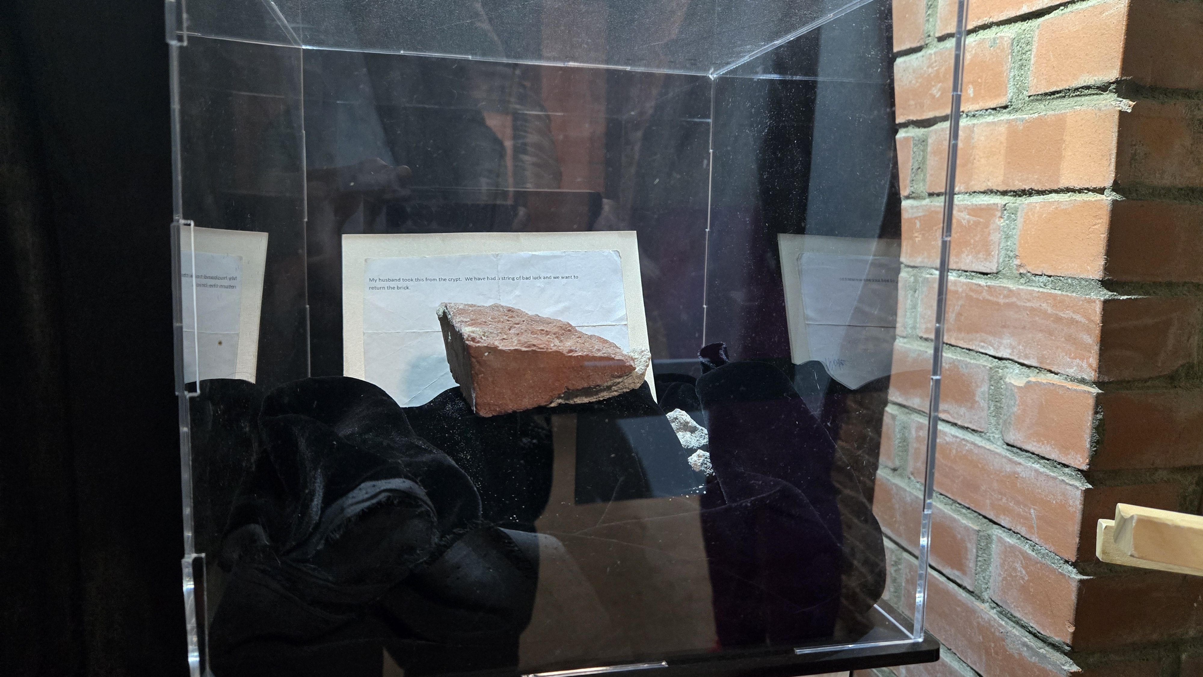A fragment of a red brick displayed inside a clear protective case with a note behind it explaining it was taken from a crypt and requests its return, set against a background of brick wall.