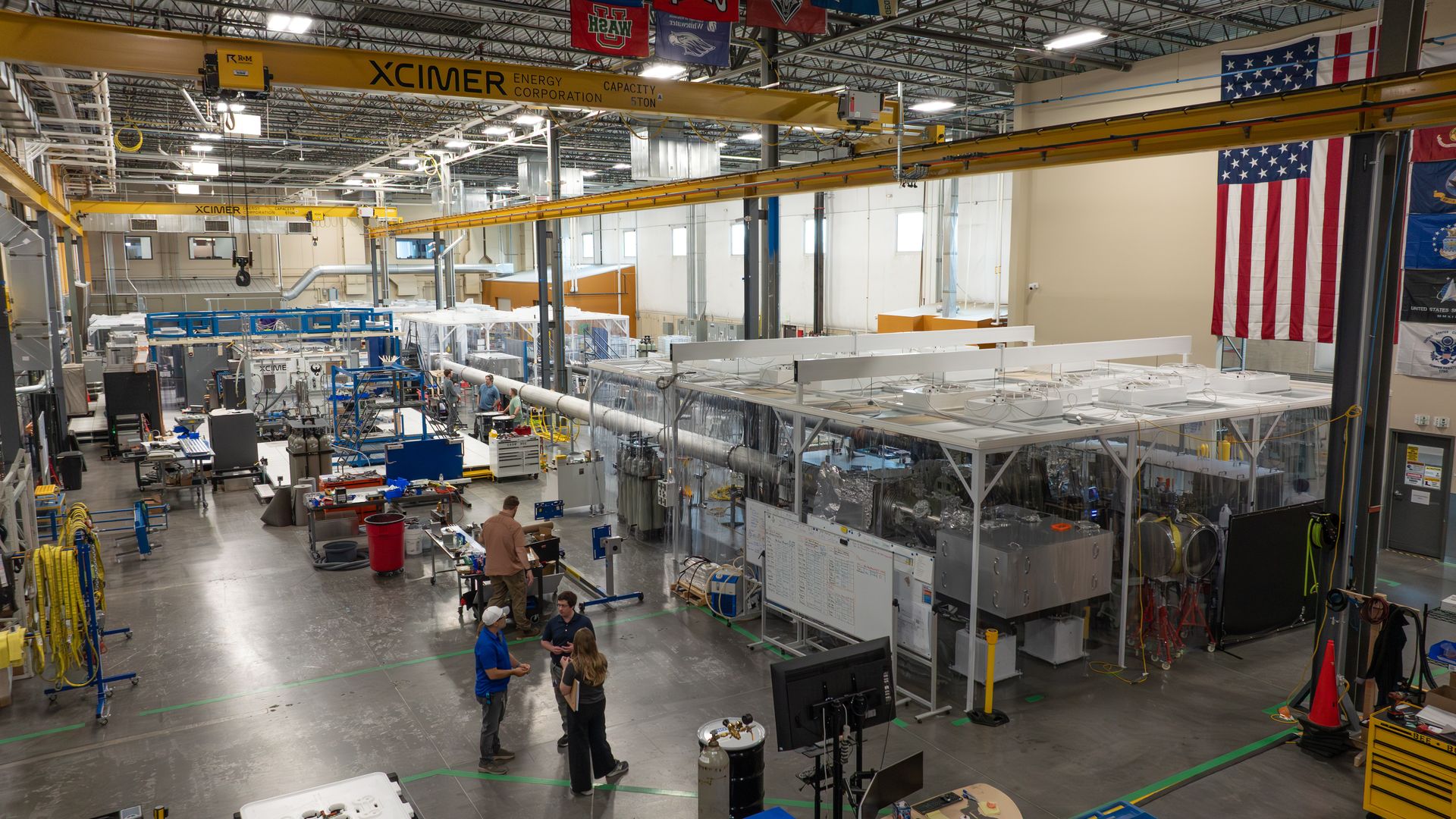 Industrial workshop with a large yellow overhead crane labeled XCIMER across the ceiling. Busy floor with blue-framed equipment, pipes, workstations, and a few workers; American flag on the wall.