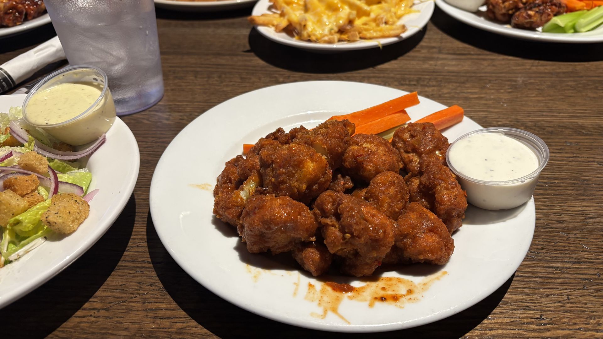 Plate of spicy buffalo cauliflower bites with carrot sticks and a cup of ranch dressing on a wooden table, with a salad and cheesy fries partially visible.