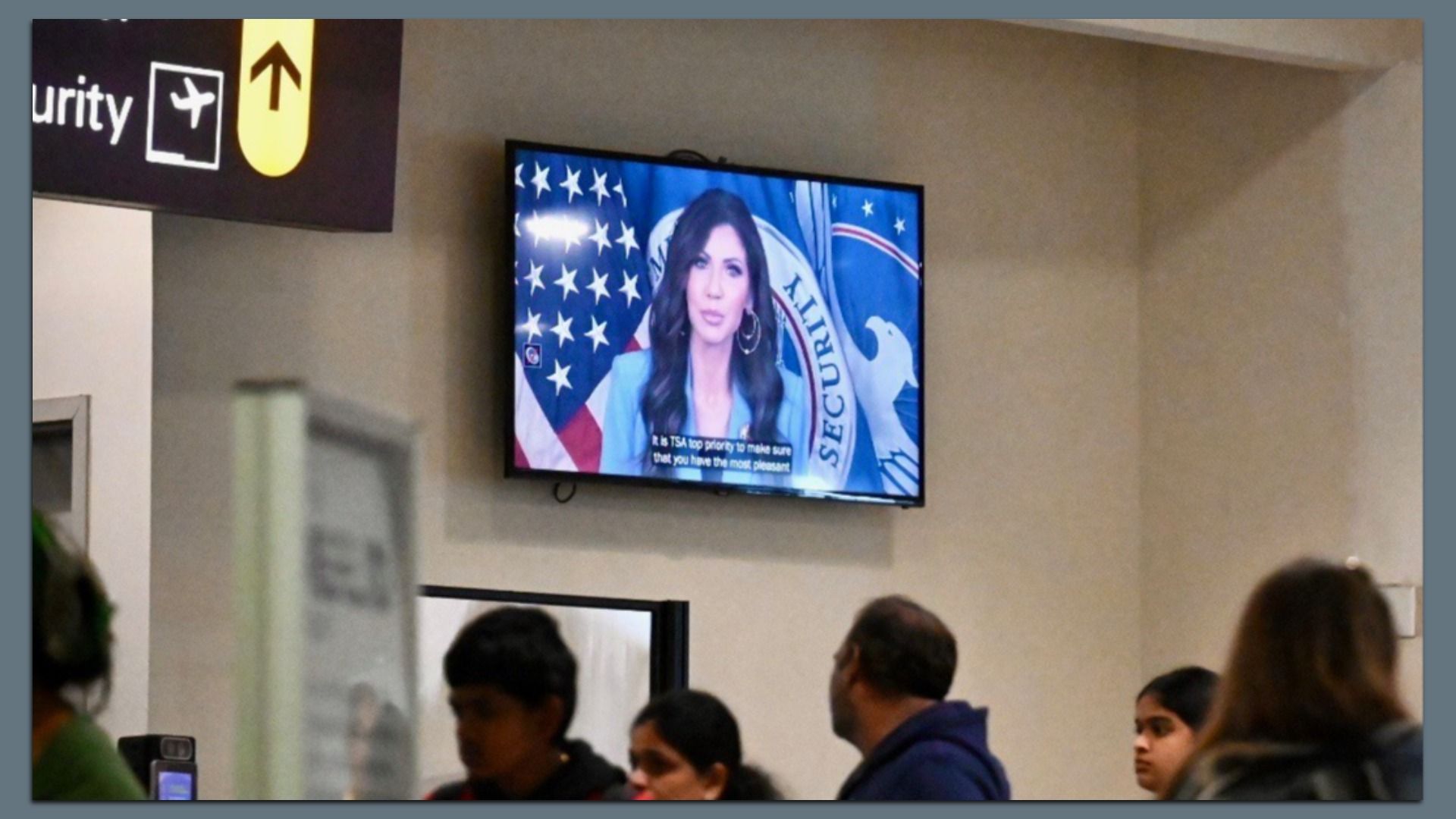 A video of Homeland Security Secretary Kristi Noem plays on a screen in a terminal with people waiting nearby. 