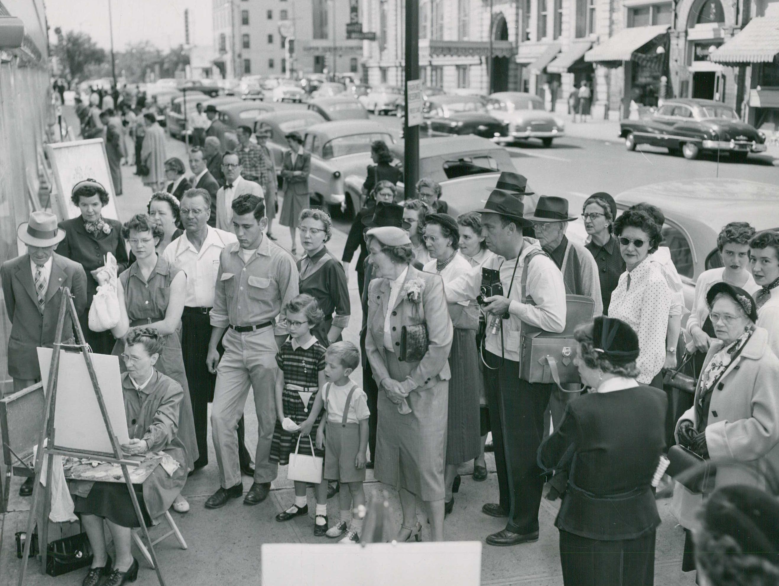 A black-and-white photo of a bunch of people gathered around a woman painting