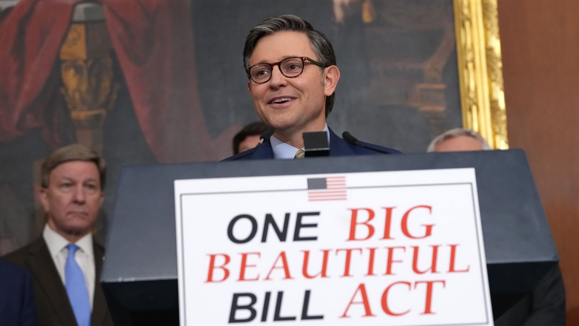 House Speaker Mike Johnson smiles as he stands behind a podium with a sign that says "ONE BIG BEAUTIFUL BILL ACT."