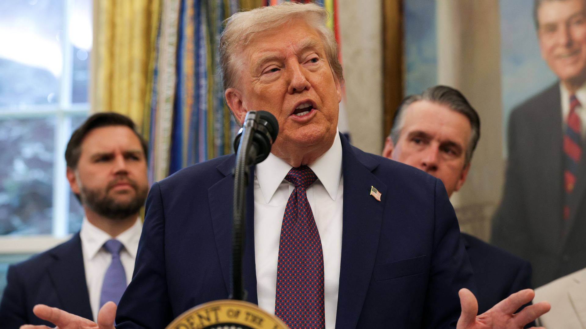 President Trump, wearing a navy jacket with a US flag pin, a white shirt and blue-maroon checkered tie raises his hands as he speaks at a podium in front of a portrait of Reagan and two men in suits.