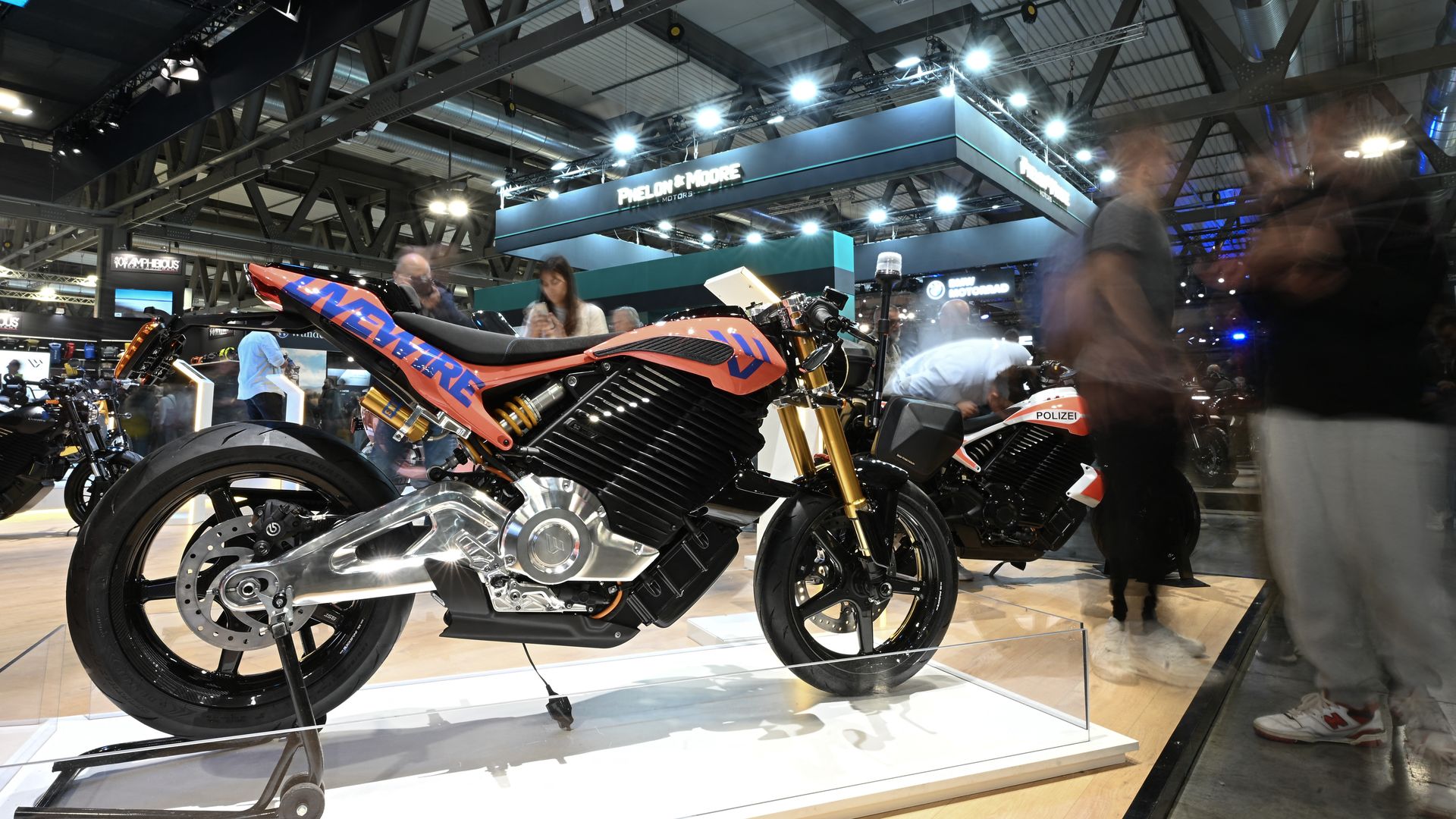 At a motorcycle show, an orange and black sport bike on a glass display stand in the foreground. Bright overhead lights, steel beams, and people walking around a busy exhibition hall.