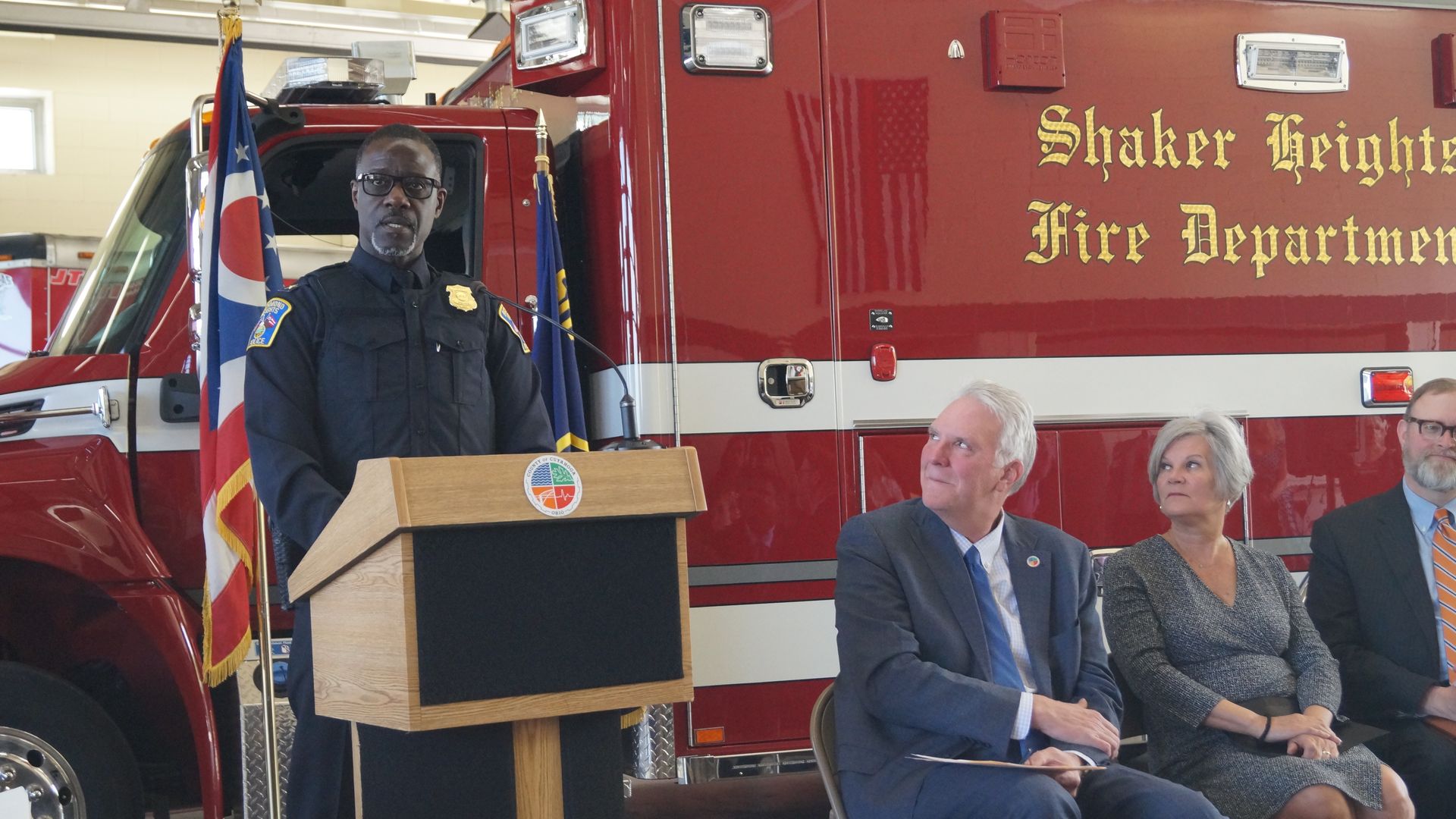 A suburban police chief standing at a lectern in front of a Shaker Heights Fire Dept truck, with seated dignitaries by his side