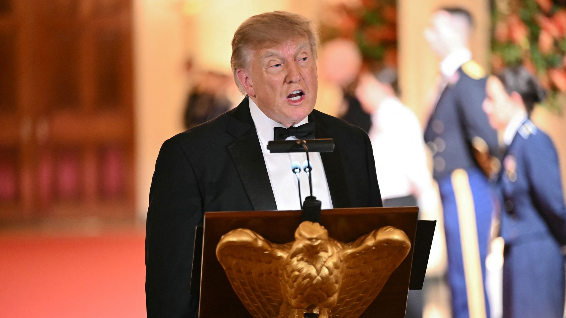 President Trump in a black tuxedo speaks at a podium with a carved eagle emblem; warm lighting and blurred background of formally dressed guests.