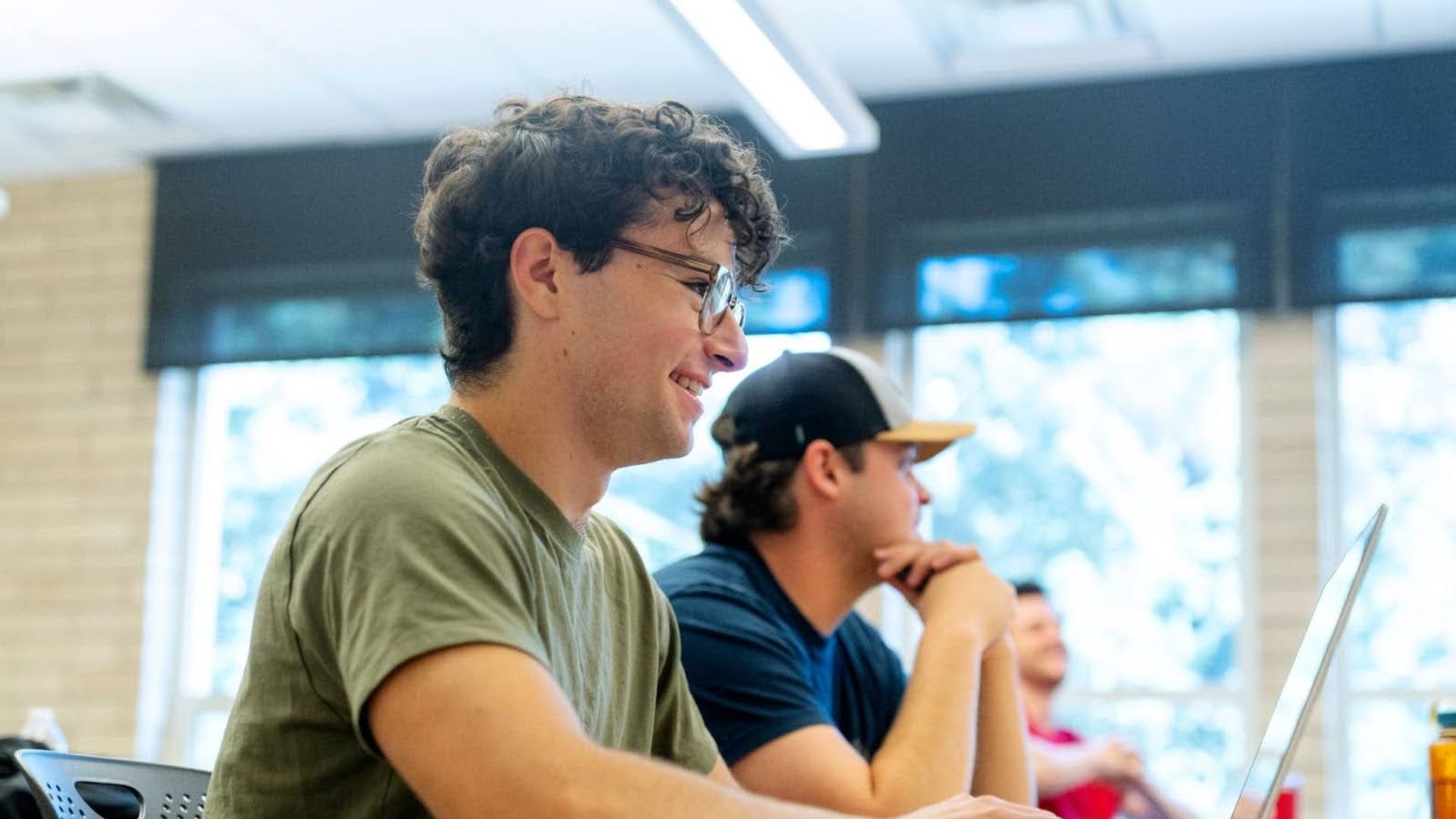 Young man with curly hair and glasses in an olive green shirt smiling while looking at a laptop, with other students seated in a classroom with large windows and natural light.