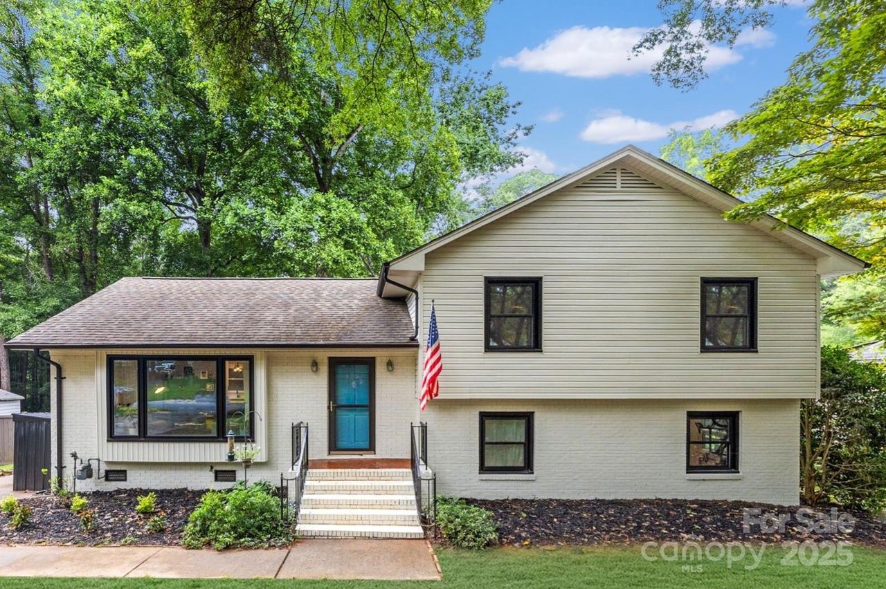 White split-level house with black window frames, blue front door, small stairs, American flag, surrounded by green trees and lawn on a sunny day.