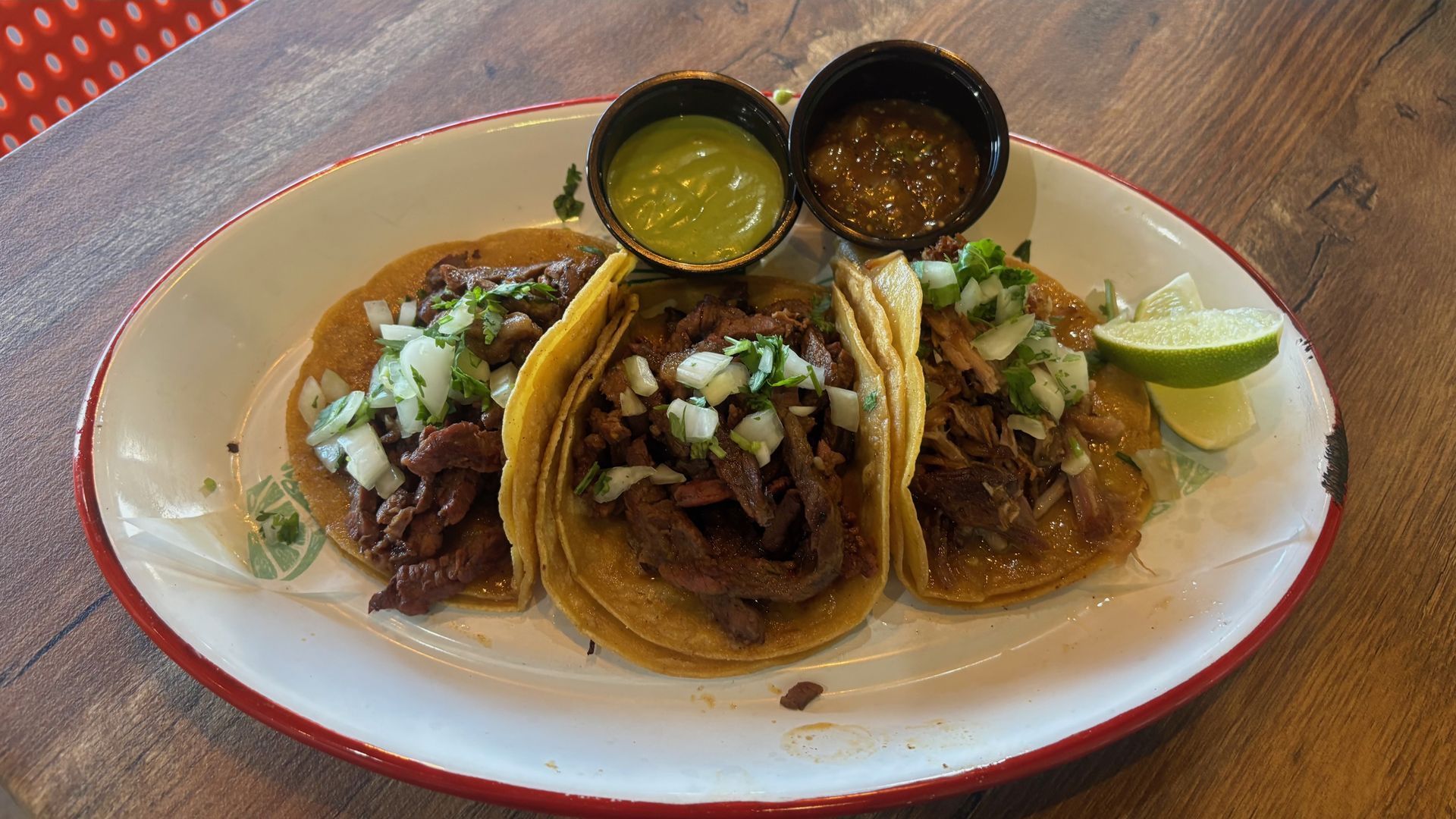 Three beef tacos plated on a white dish with chopped onions and cilantro, served with green avocado sauce and brown salsa in small cups, a lime wedge on the side, on a wooden table.