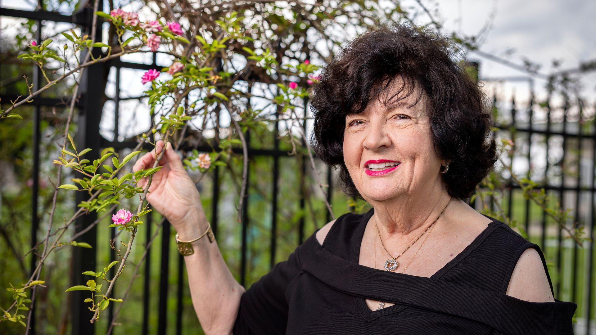 Smiling older woman with dark curly hair in a black dress stands by a flower-covered trellis, holding pink blossoms, with a cloudy sky and black fence in the background.