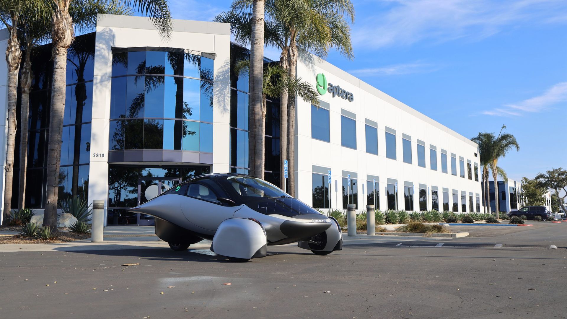 Sleek, futuristic three-wheeled silver and black vehicle parked outside a modern white office building with large blue-tinted windows and palm trees under a clear blue sky.