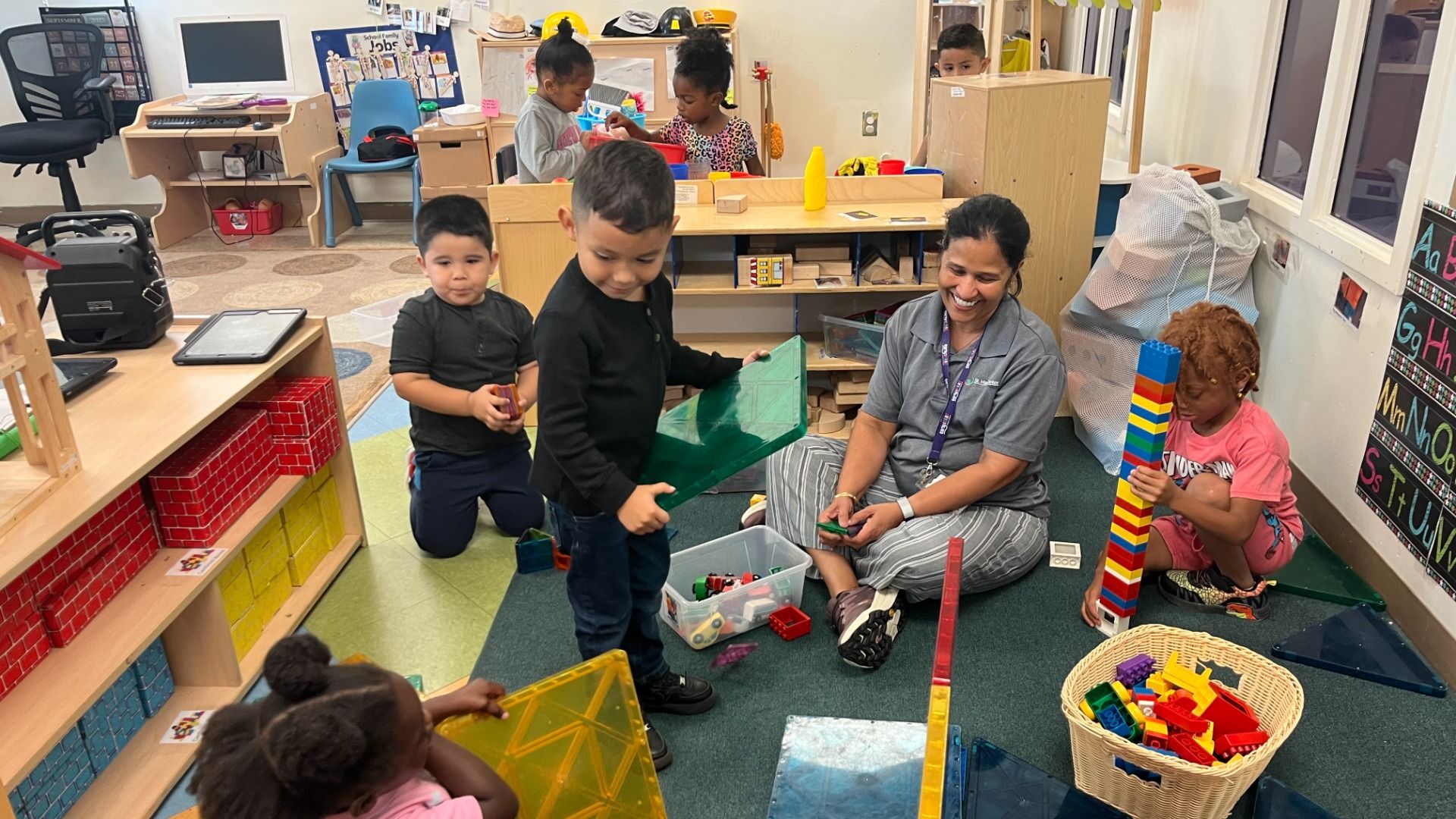 A classroom scene with children and a teacher playing with colorful building blocks and geometric shapes. The teacher is smiling, and kids are engaged in building and sorting toys.