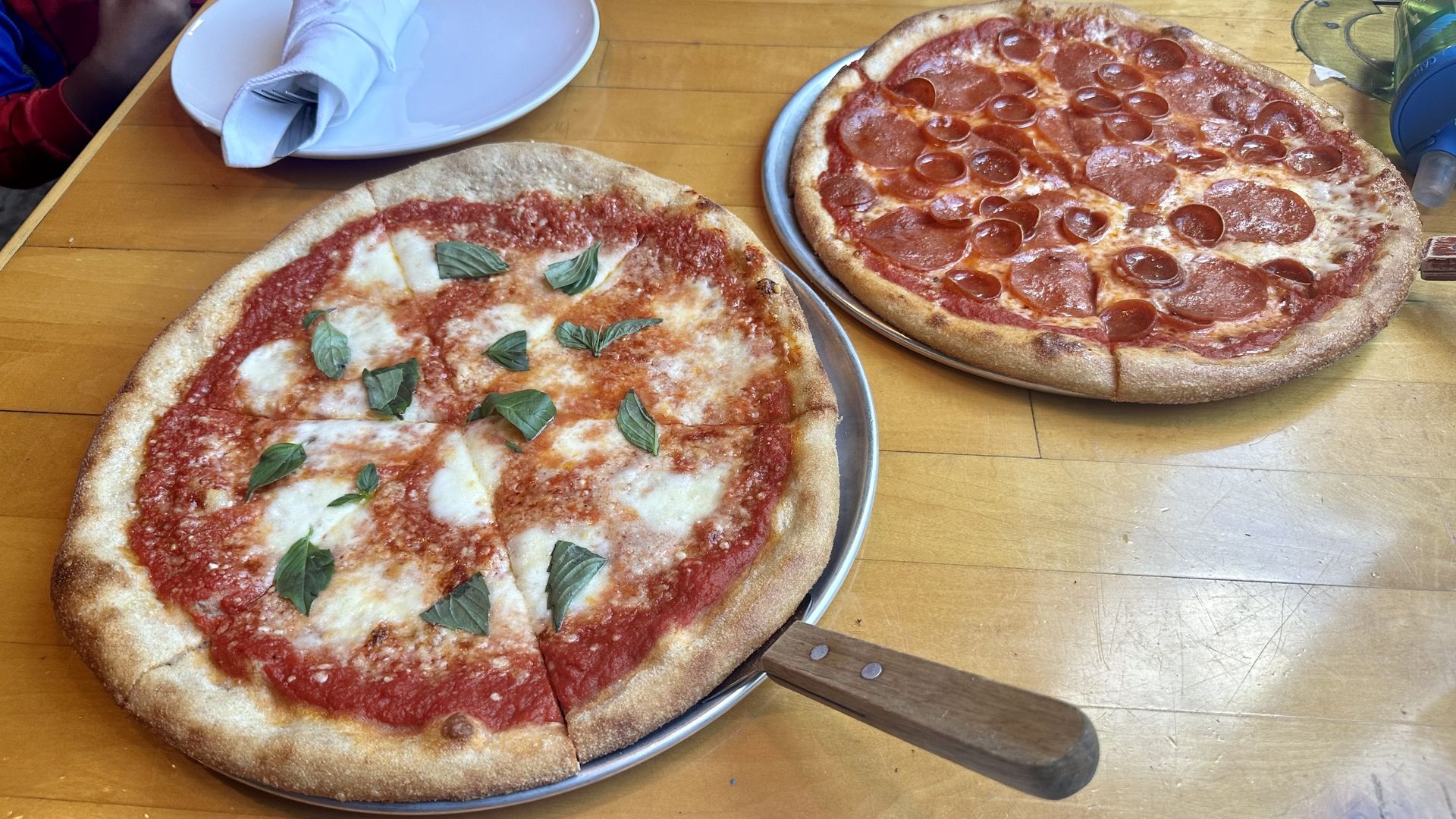 Two pizzas are shown on a table: a margherita-style cheese pizza at left foreground, and a pepperoni in right background. The cheese pizza has a pizza serving utensil underneath it.