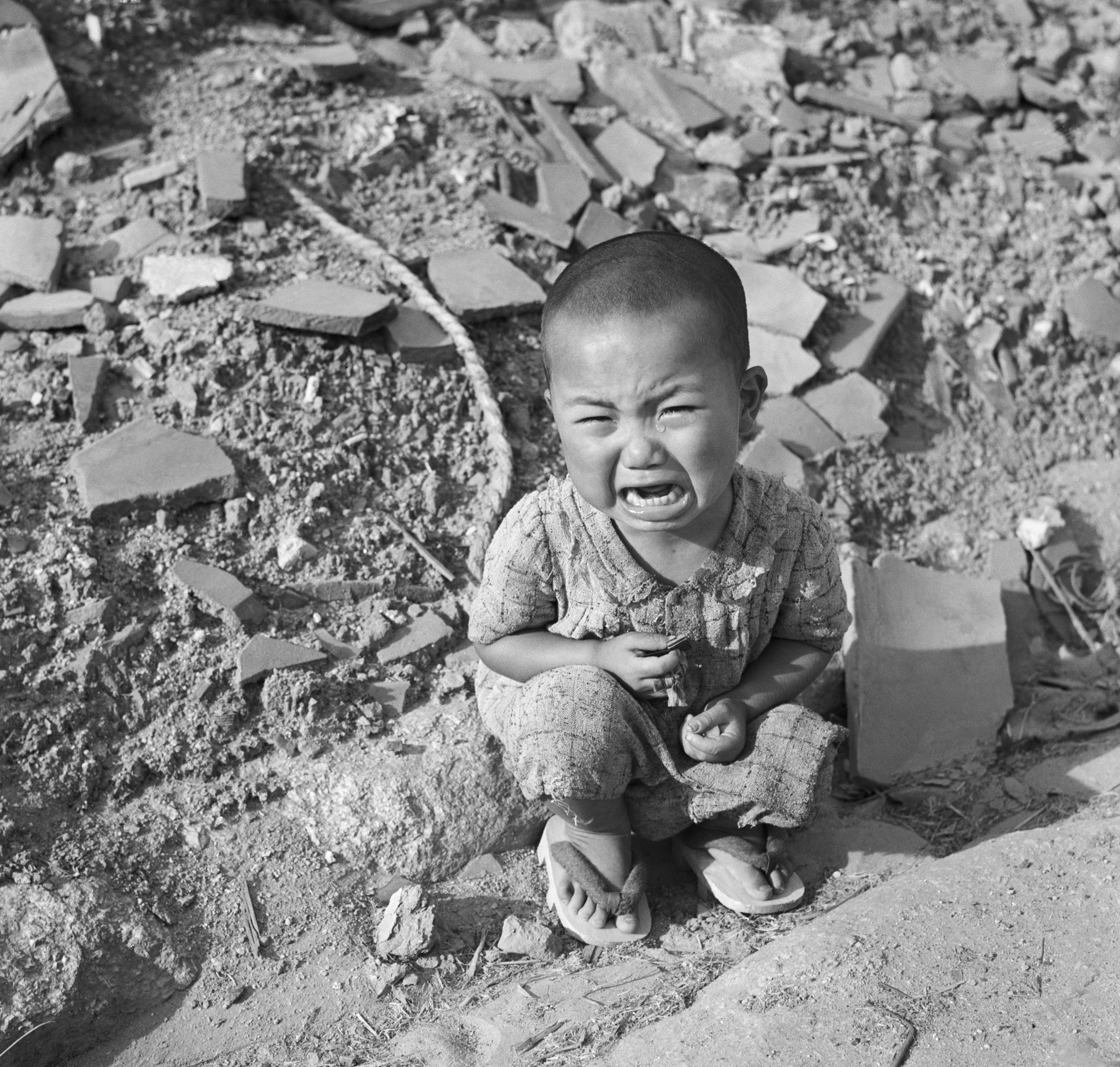 Black and white image of a young child crying while crouching on a rough ground strewn with broken bricks and debris in Hiroshima, wearing a patterned outfit and sandals.