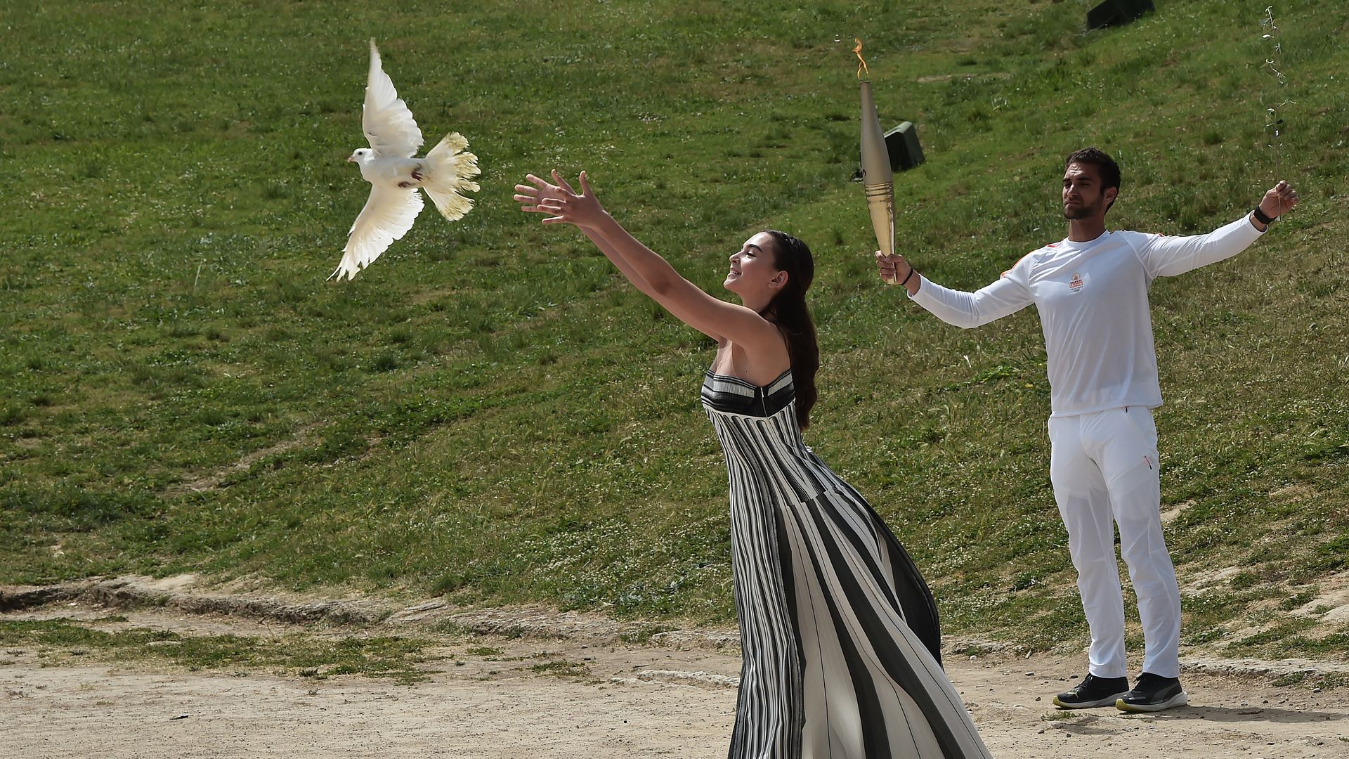 Two people are photographed during the torch lighting ceremony. A woman, dressed in a black and white sleeveless dress, is releasing a bird. A man, dressed in all white, holds the torch behind her. 
