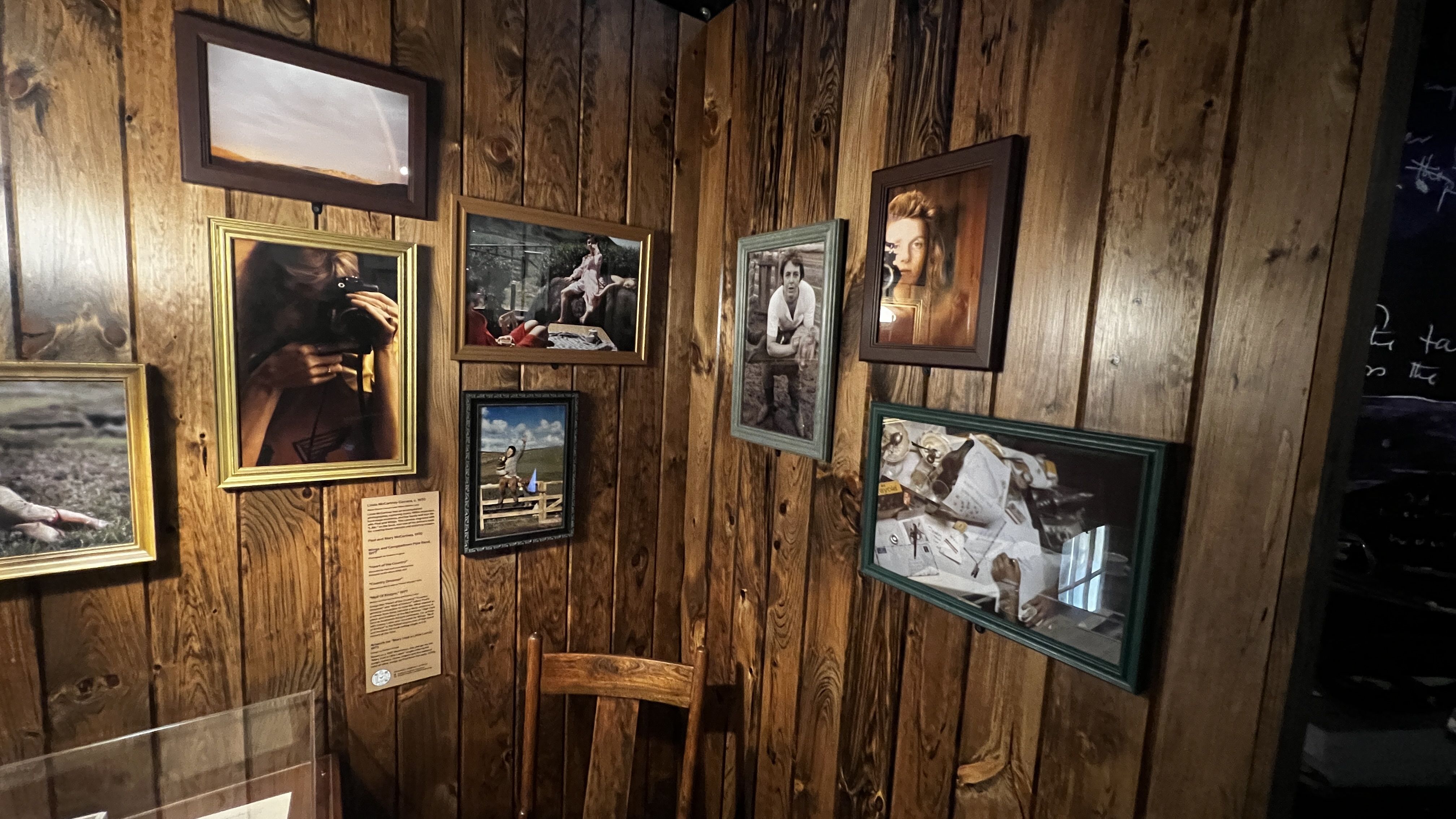 Wooden wall with a collage of framed photos in gold, green, and brown frames; portraits and landscapes hang at odd angles, with a wooden chair and a glass display case in front.