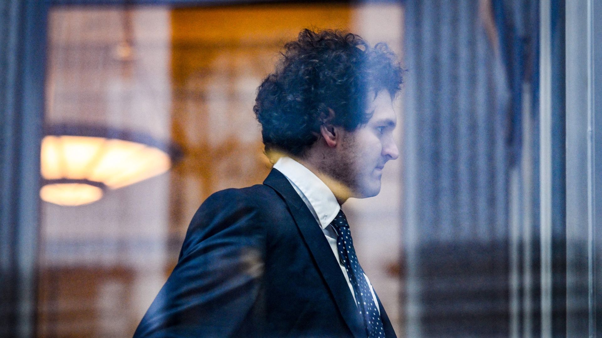 A man in a suit is seen through a window, walking with a serious look on his face.