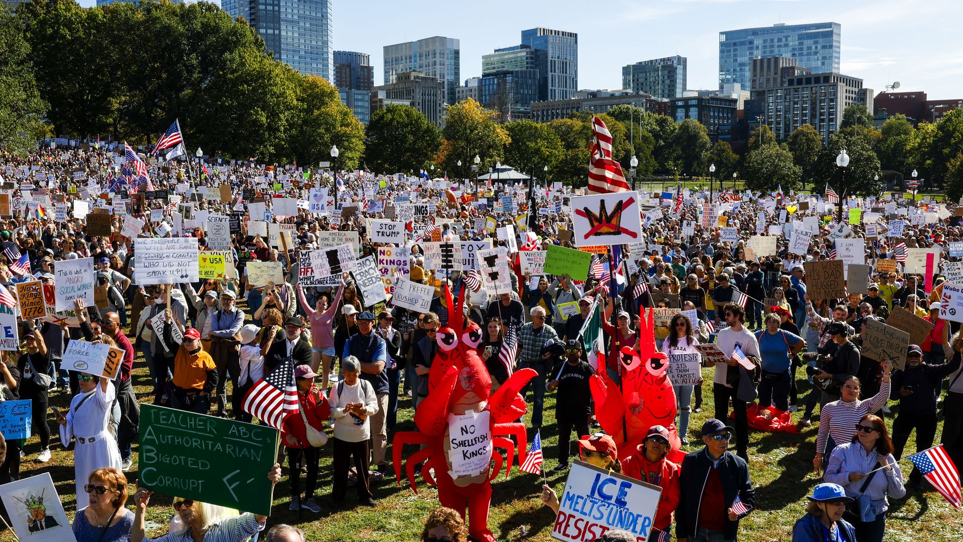 A large crowd of protesters fills a city park, waving American flags and holding handmade signs; glass skyscrapers rise behind trees in the bright day, with some people wearing red lobster costumes.