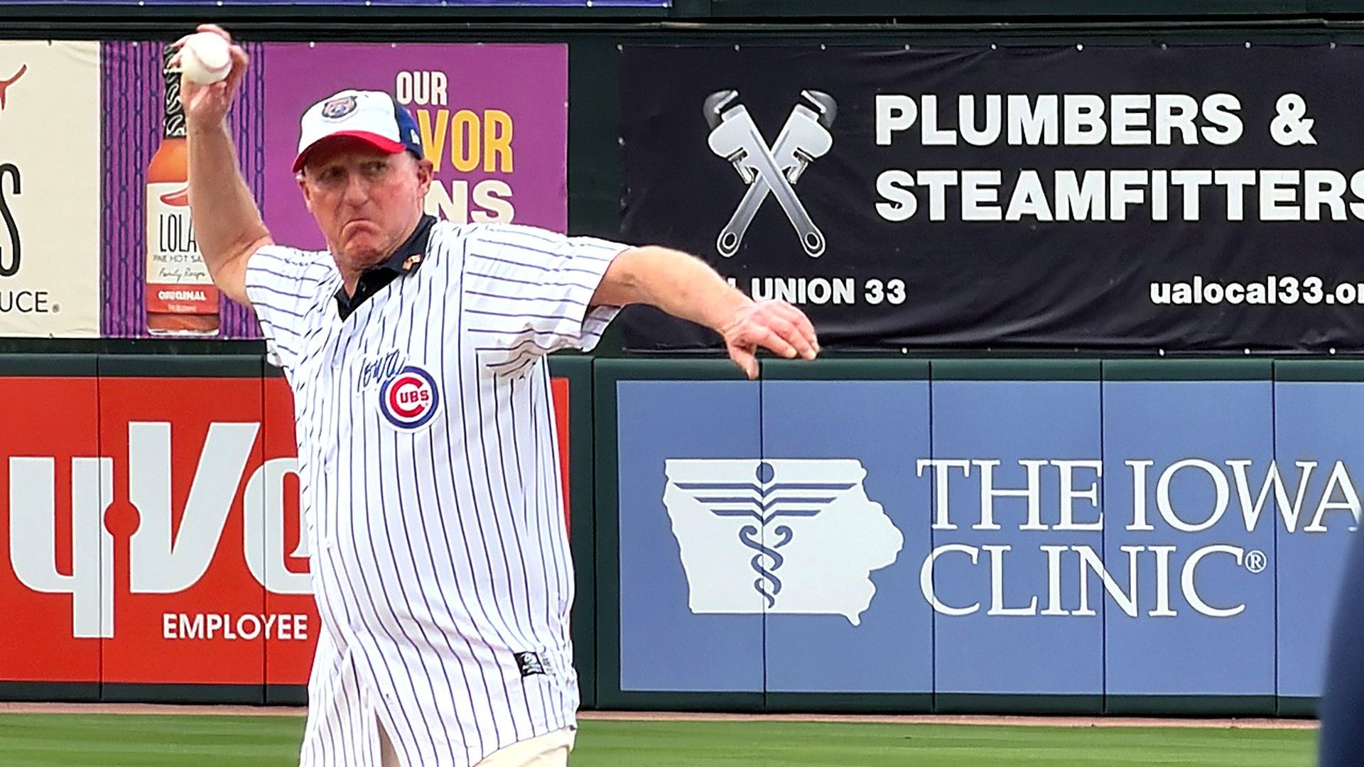 Chicago Cubs pitcher in a white pinstriped uniform and a red-blue cap throws a pitch on the field. The outfield wall banners read "THE IOWA CLINIC" and "PLUMBERS & STEAMFITTERS."