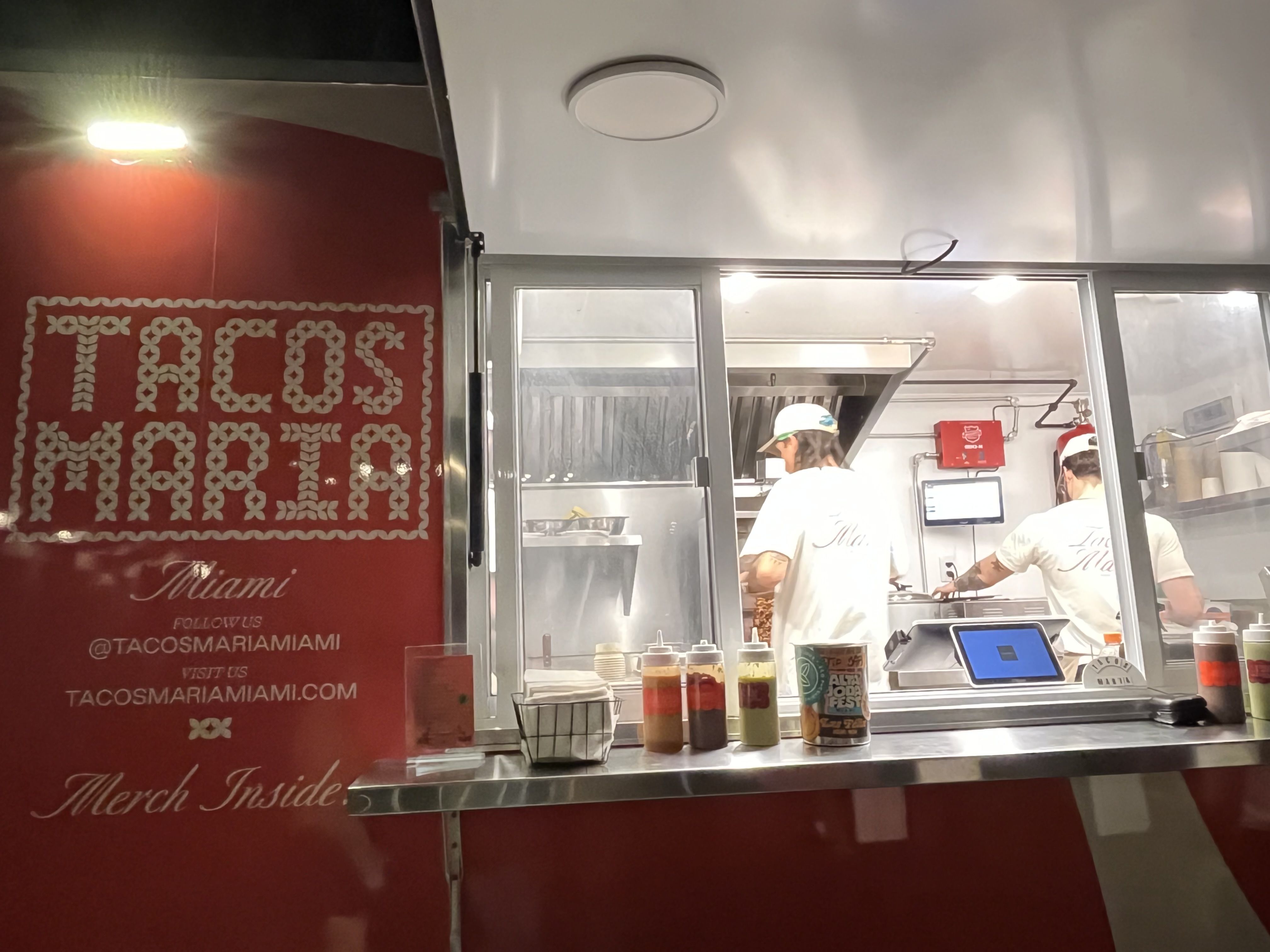 Night view of Tacos Maria food truck with red exterior, two workers inside preparing food, sauces on counter, and signage with social media and website details.