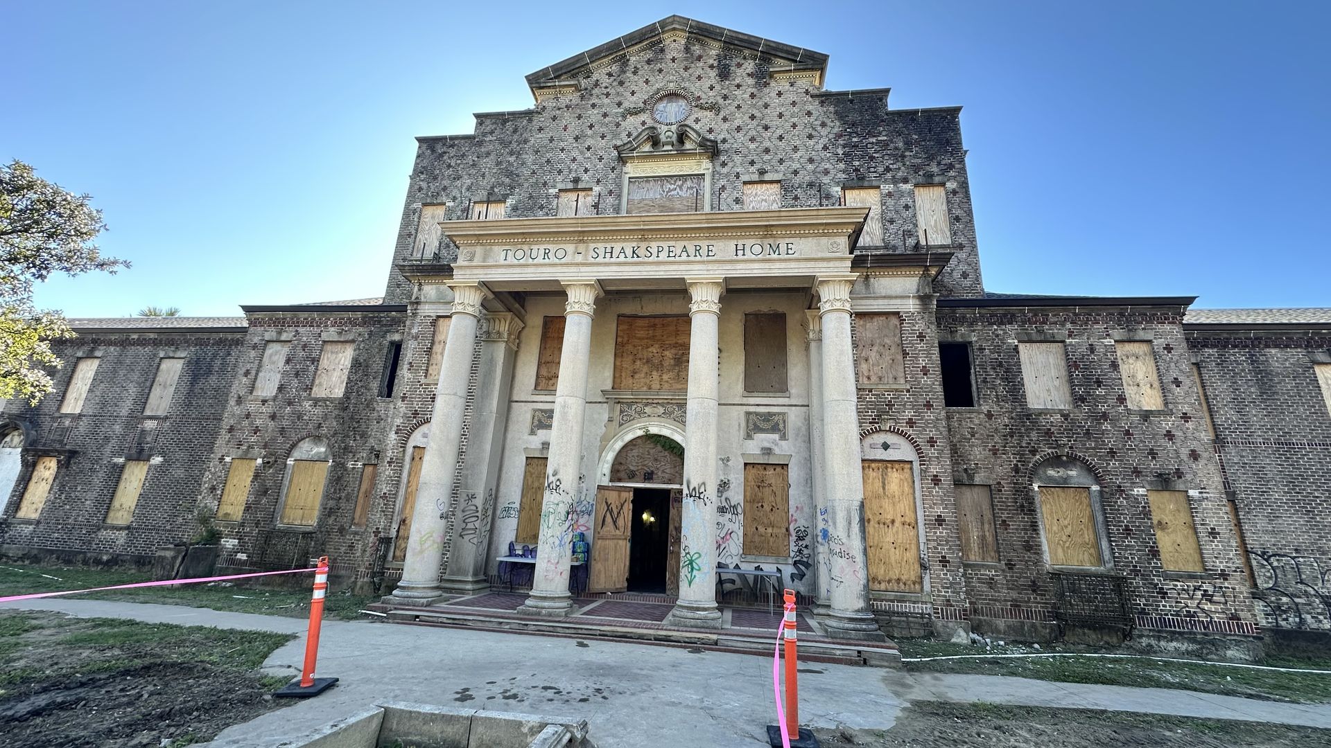 The Touro-Shakspeare Home, an old brick building with boarded-up windows and graffiti on columns, under a clear blue sky with a walkway and orange construction cones in front.