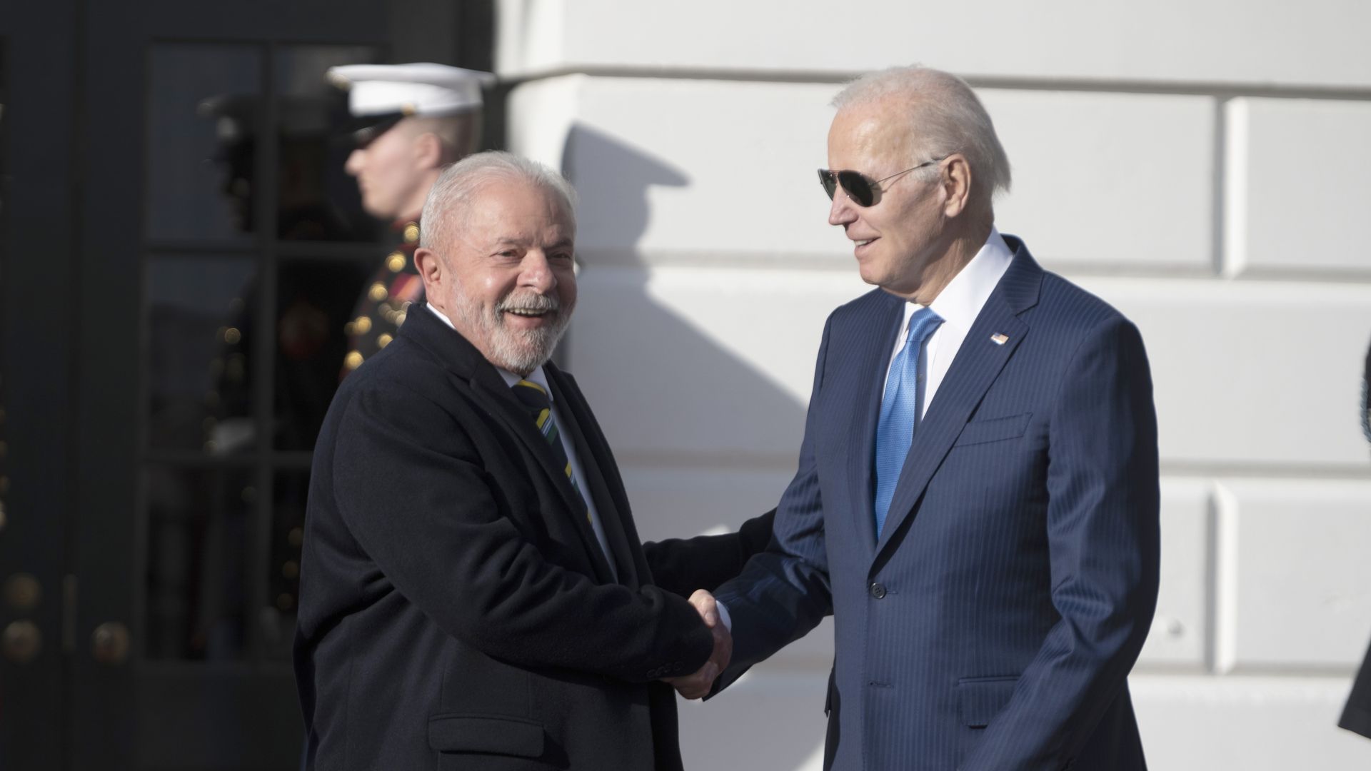 Brazil President Luiz Inácio Lula da Silva, wearing a black coat, smiles as he shakes hands with President Biden, who is in a navy blue suit and sunglasses. They are outside the White House. 