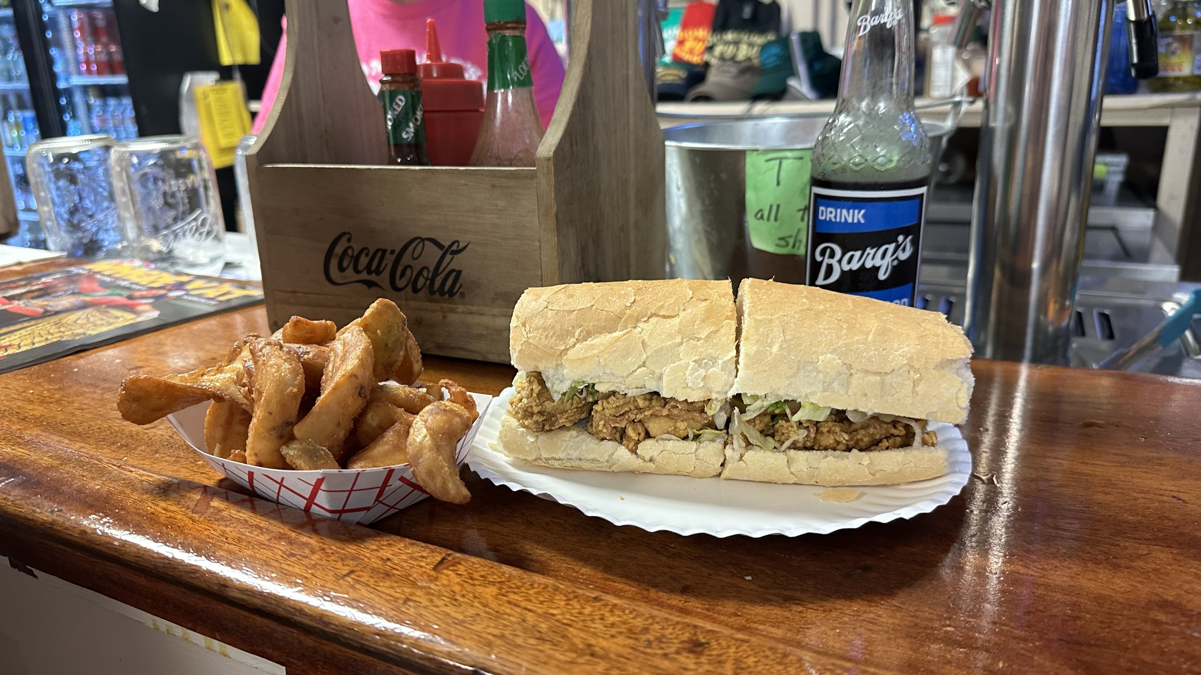 An oyster poboy sits on a white paper plate next to a paper basket of fries on a bartop. A root beer is seen in the background.