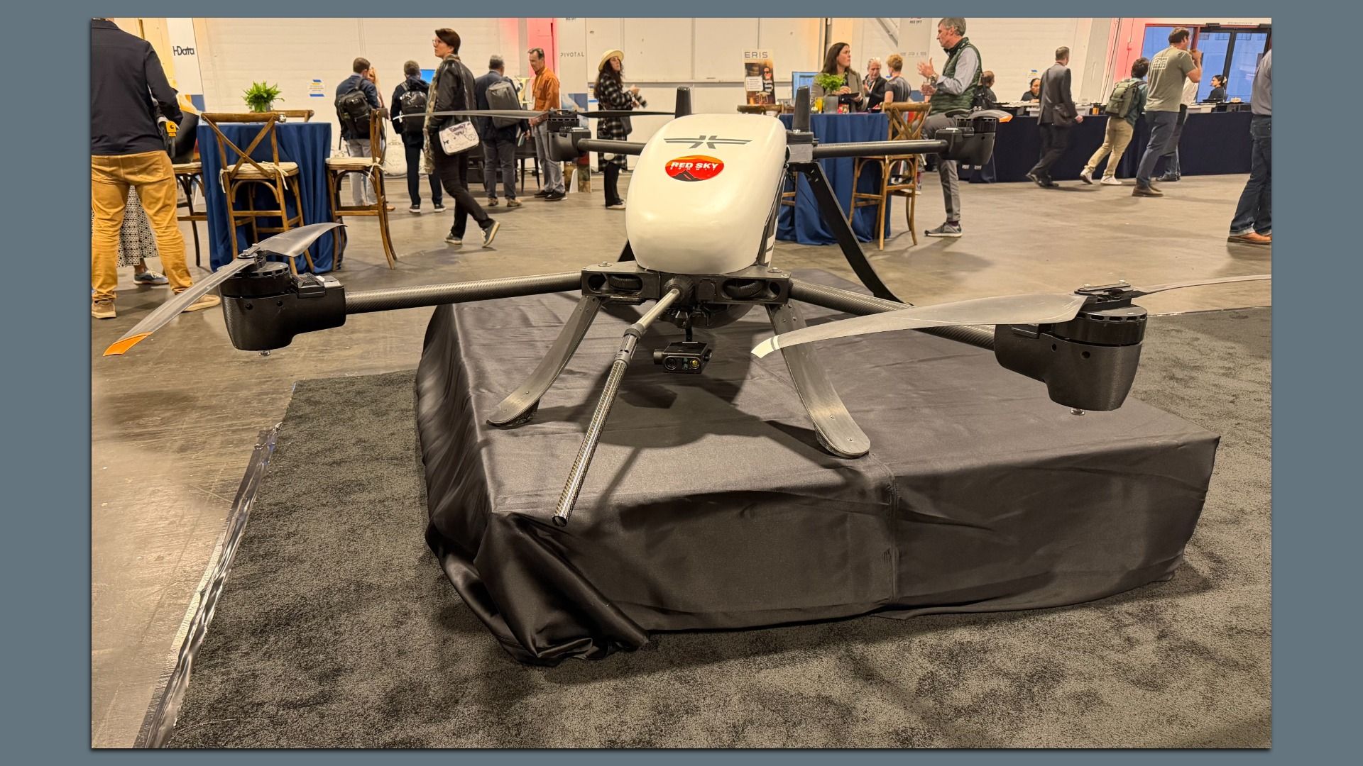 Large white and black drone with four propellers displayed on a black-covered platform at an indoor event with people and tables in the background.