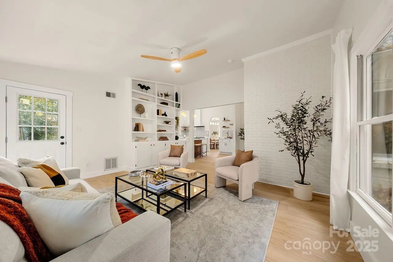 Bright living room with white walls and ceiling, light wood floor, gray rug, beige sofa with pillows, two cushioned chairs, glass coffee table, built-in shelves, ceiling fan, and potted plant by window.