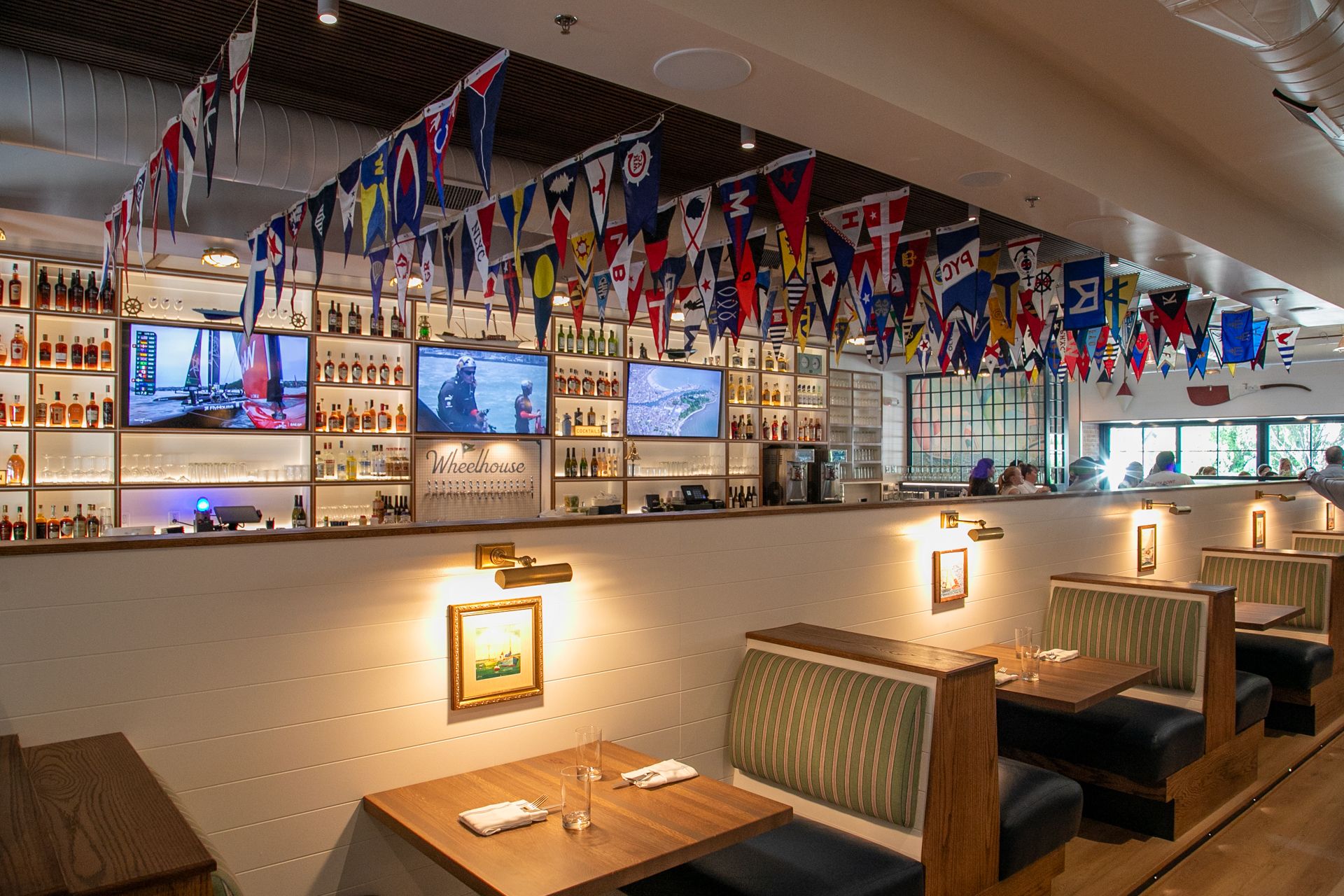 Spacious, cozy bar with white paneled walls, wooden booths with green-striped cushions, and a ceiling of colorful nautical pennant flags. Bottles line back shelves and TVs show sailing scenes.