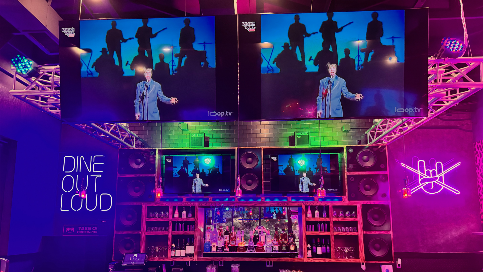 Colorful nightclub bar with two large screens showing a man in a light blue suit singing; purple lighting, stage truss, neon DINE OUT LOUD, and a stocked bar with tufted stools.