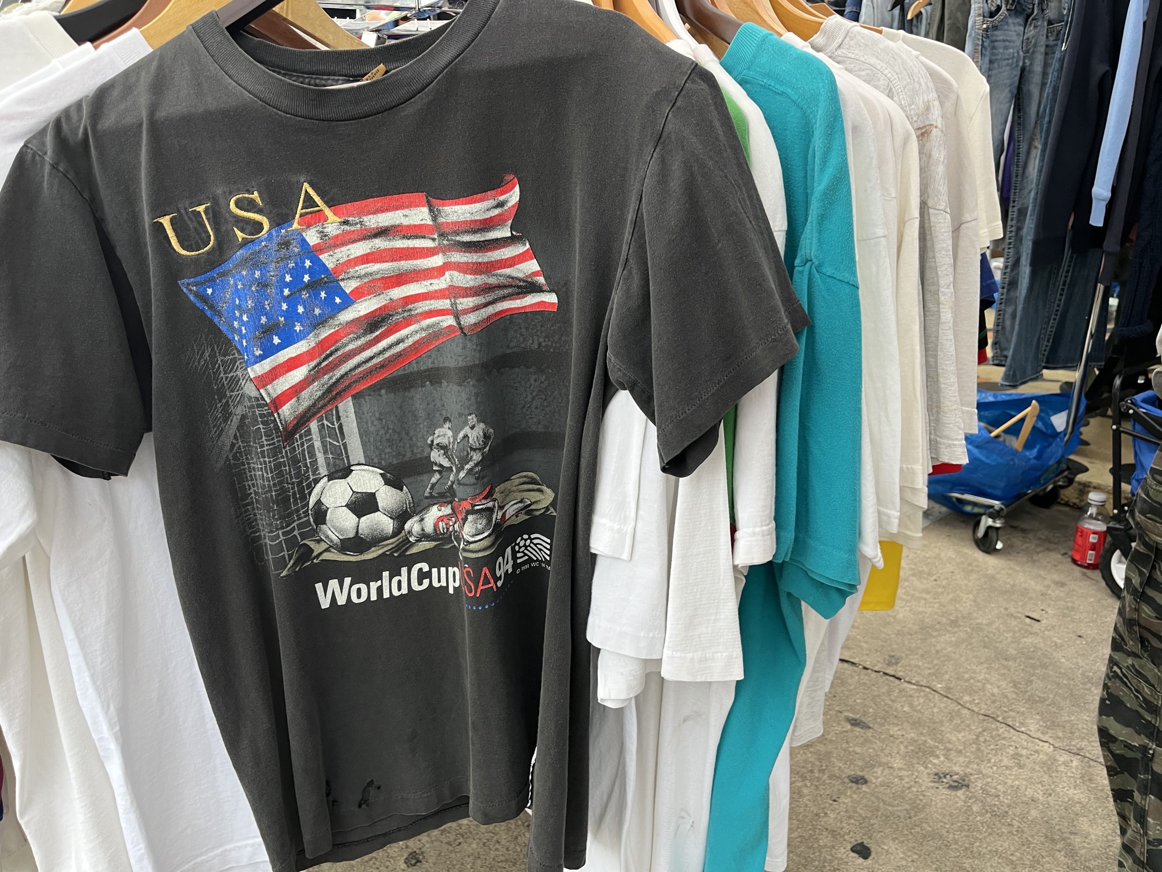 Black vintage T-shirt on a clothing rack featuring a waving USA flag, a soccer ball, and the text "World Cup USA 94" on it, surrounded by white, teal, and other shirts in a thrift store.