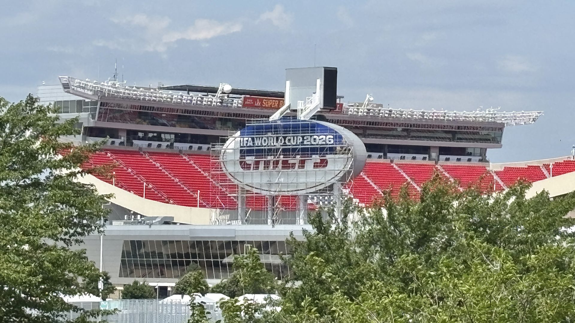 Red-seated stadium with a modern circular glass sign reading "FIFA World Cup 2026"; blue sky above and trees in the foreground.
